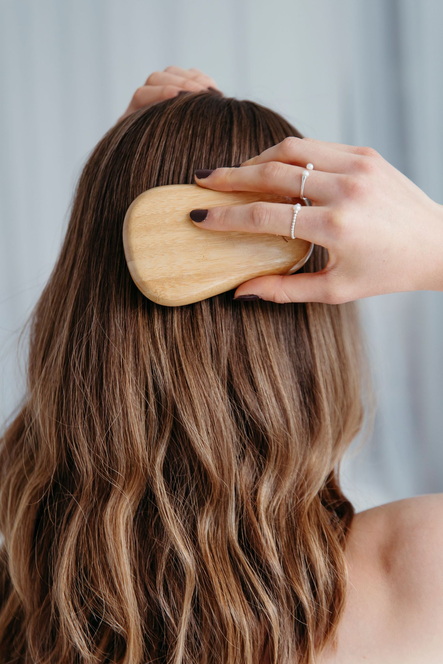 Wooden hair brush being used to style wavy brown hair against light background.