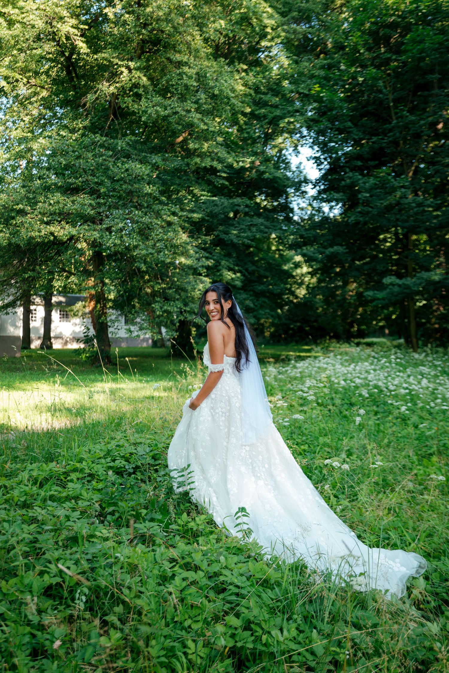 A flowing white wedding dress trails behind as someone walks through lush green summer gardens.