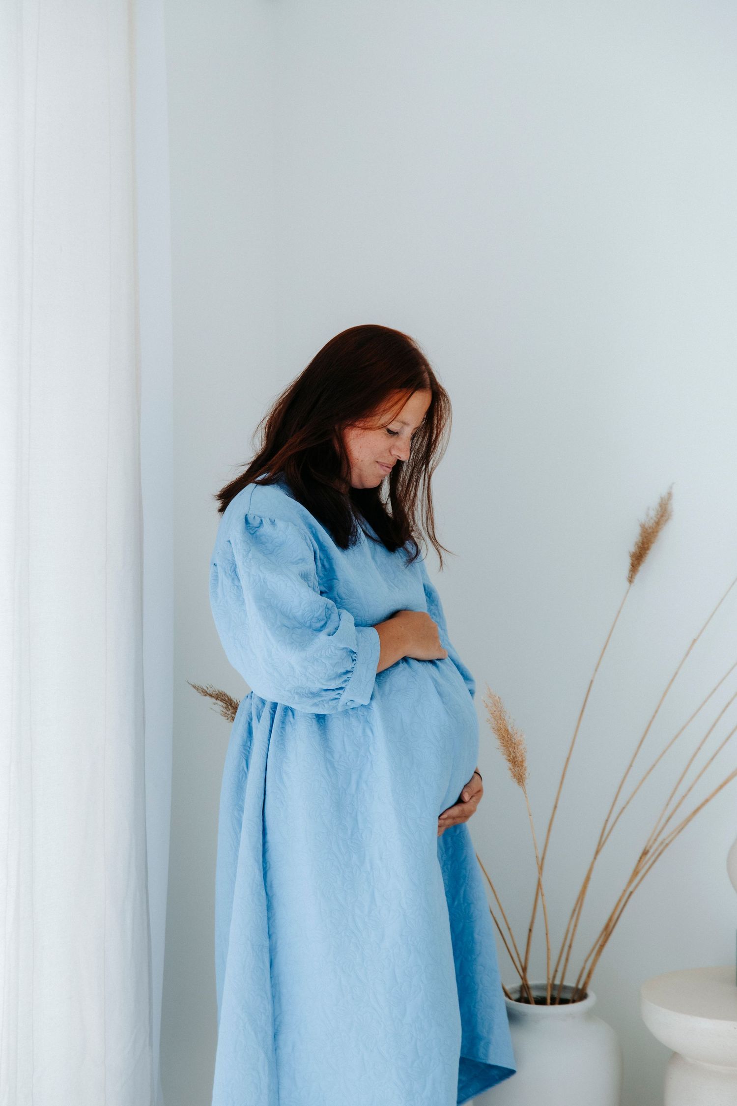Person in flowing light blue dress poses against white backdrop with dried pampas grass.