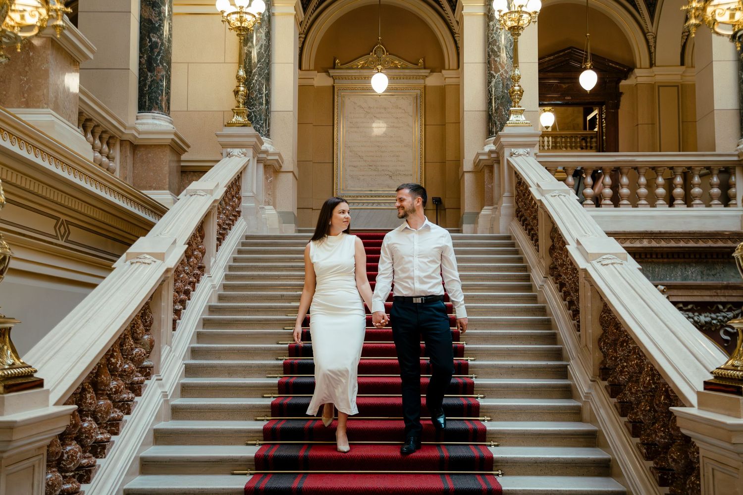 Couple descends grand marble staircase with red carpet in elegant historic building.