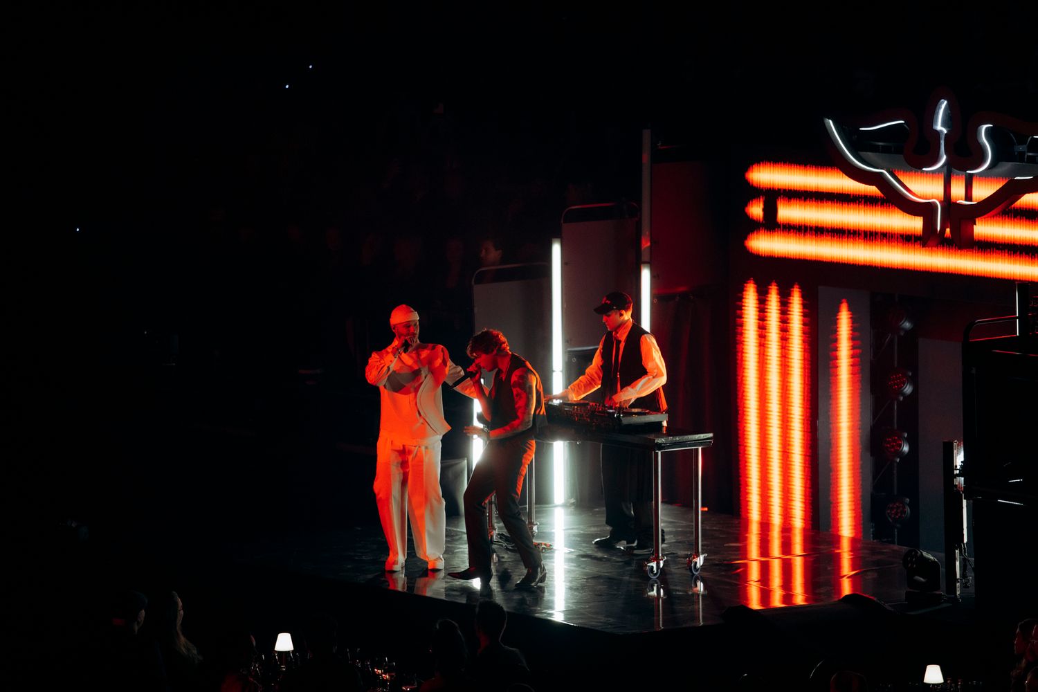 Silhouetted figures perform on a dark stage with glowing orange light bars in the background.