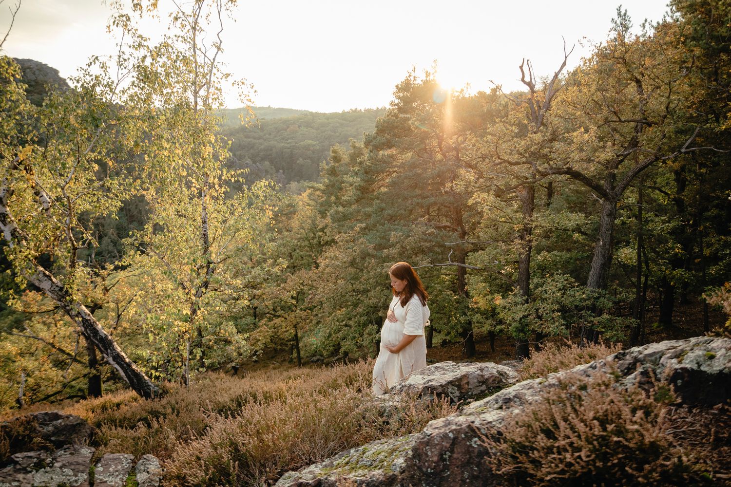 Expectant mother in white dress standing on cliff edge during golden hour.