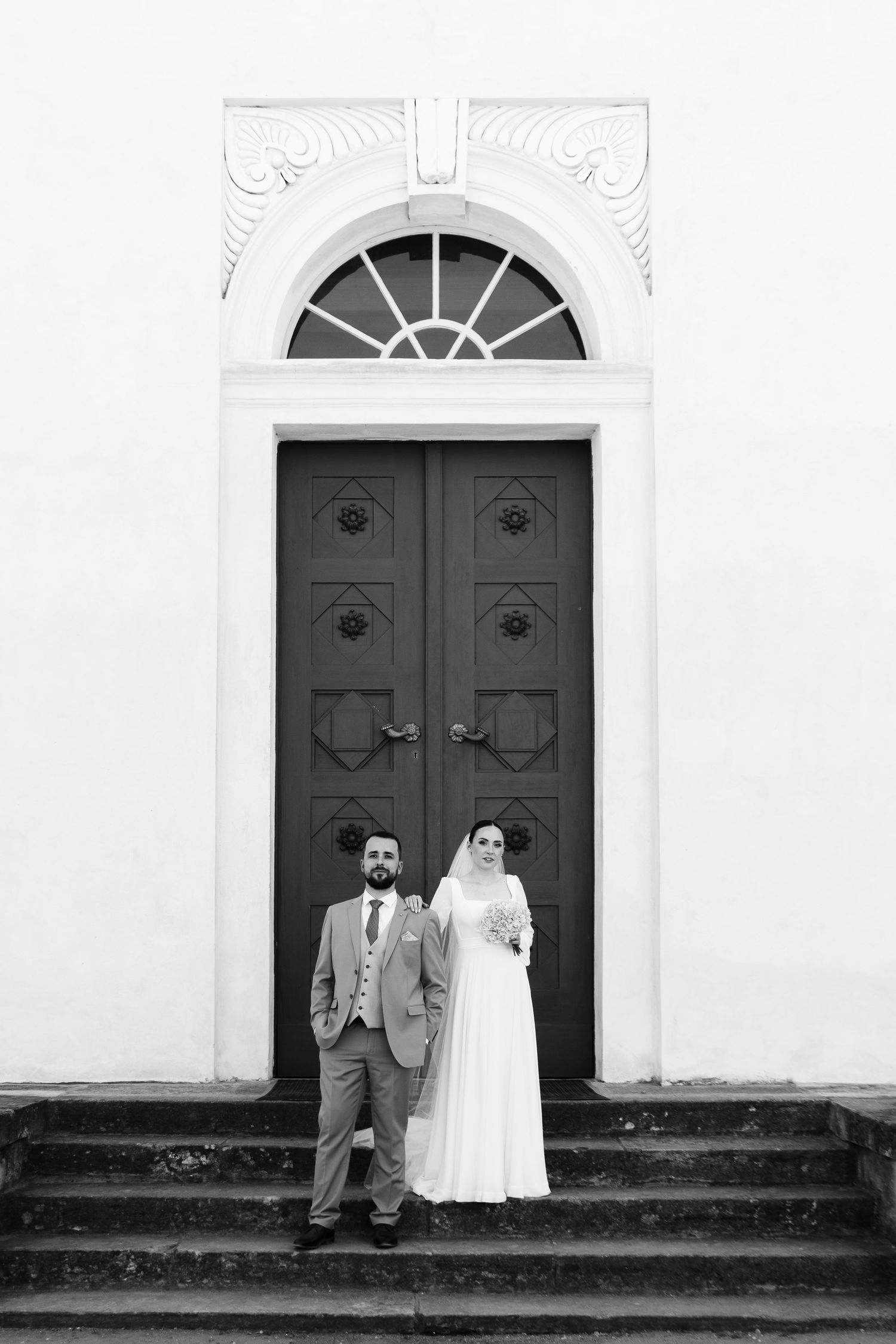 Black and white wedding photo of bride and groom standing before ornate church doors on stone steps.