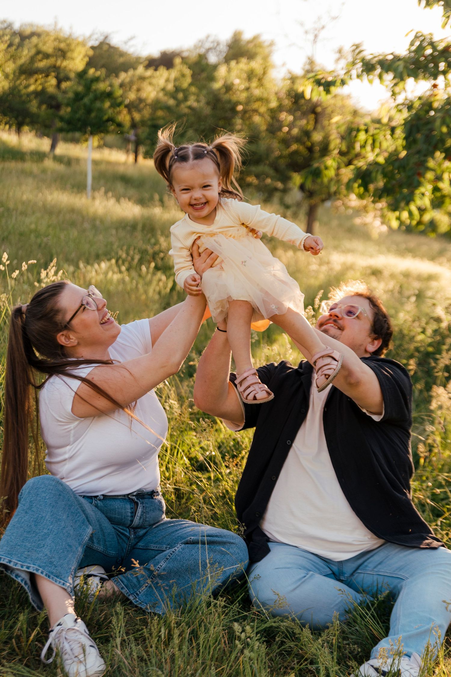 Happy family sitting on grass, lifting laughing toddler girl in yellow dress outdoors in sunny green park.