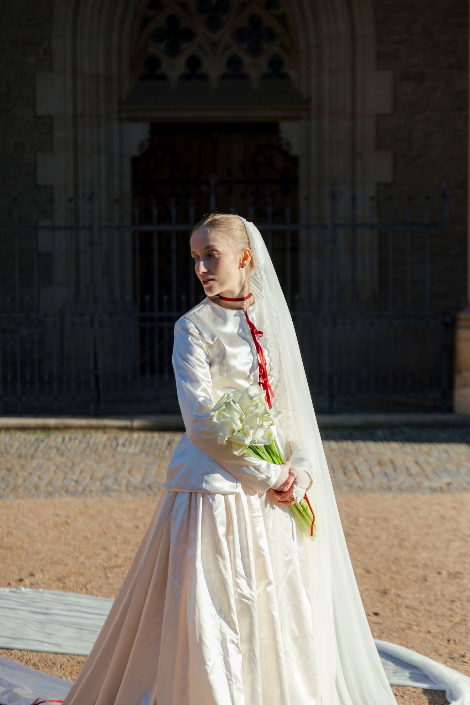 A bride in an elegant white wedding gown and veil poses outside a historic stone church.