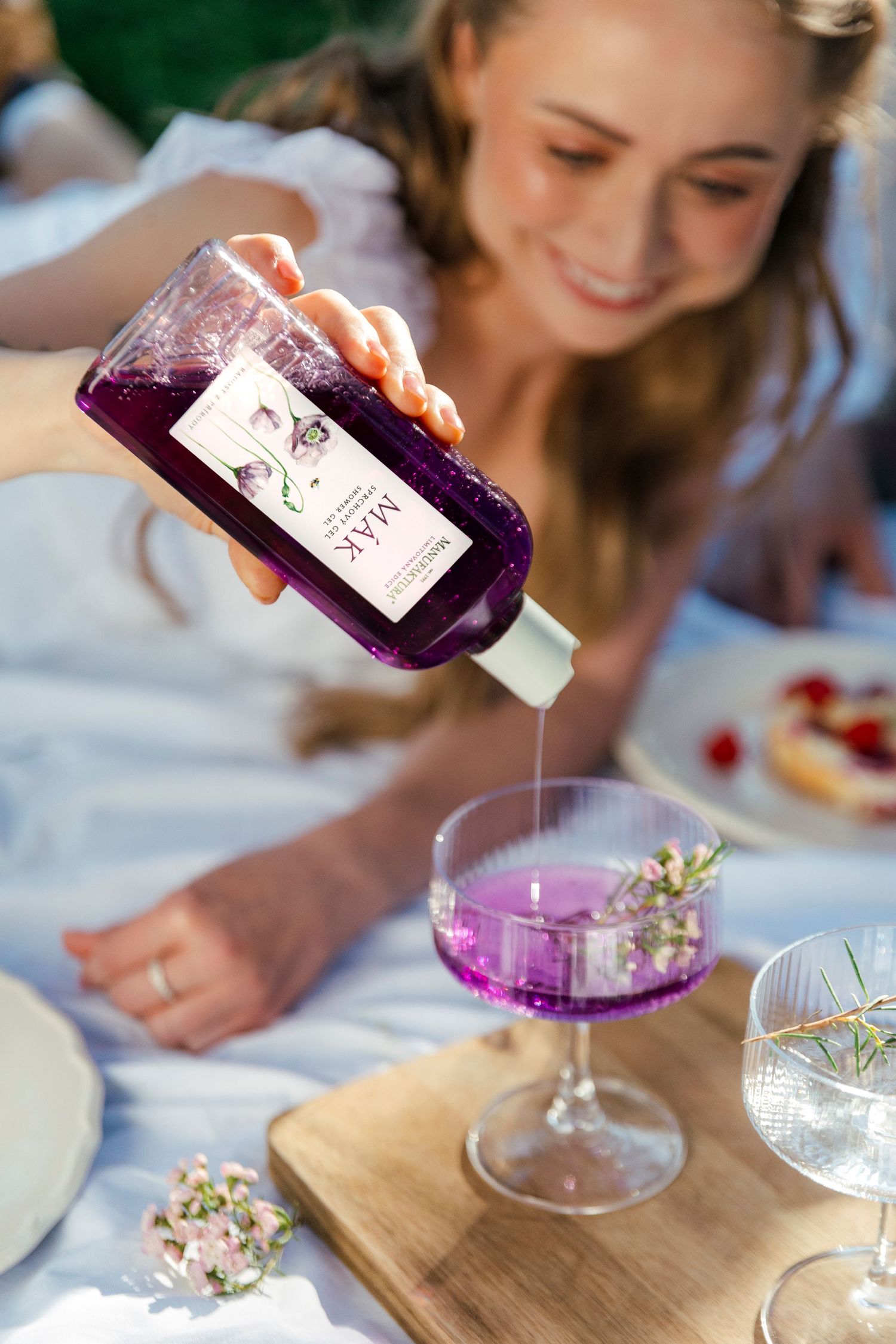 Woman pouring purple lavender syrup from a MAK bottle into an elegant cocktail glass at a picnic setting.