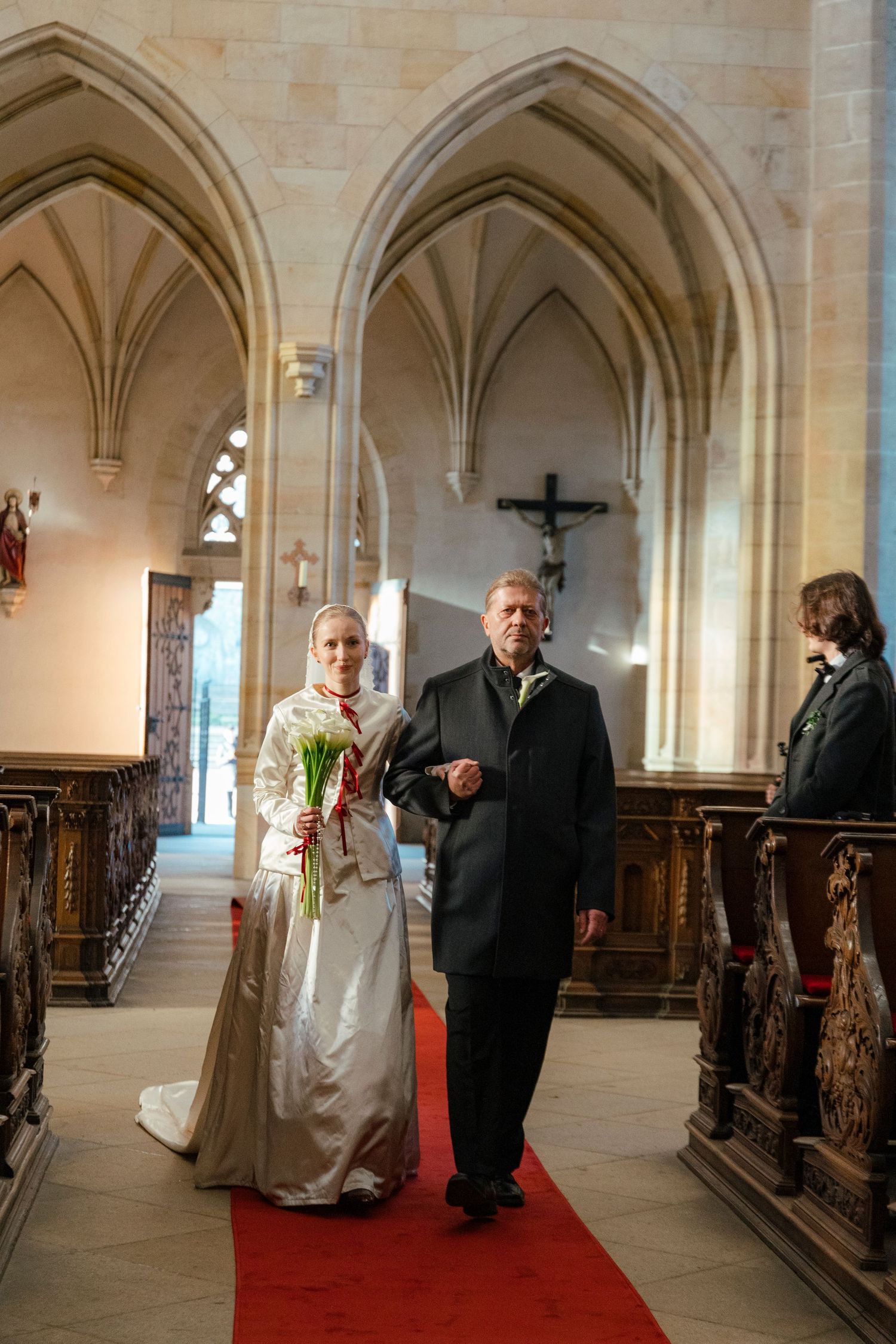 Gothic stone arches line a church hallway with wooden pews and a red carpet runner.