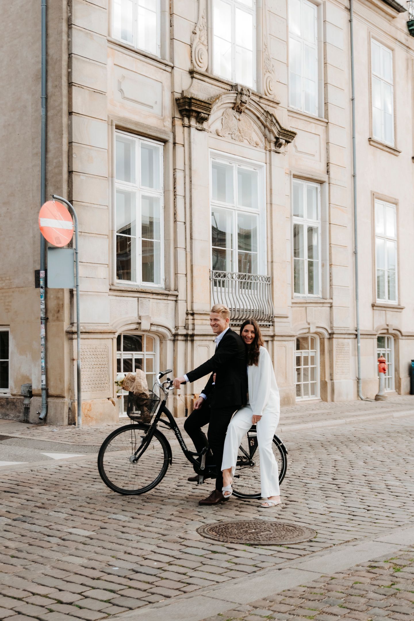 Couple posing with a vintage bicycle on cobblestone street near historic building.