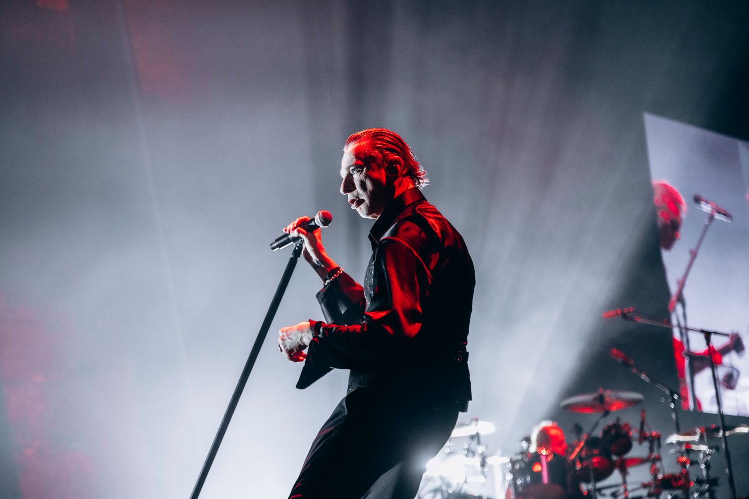 Concert performer in red lighting sings on stage with atmospheric fog effects during live music performance.