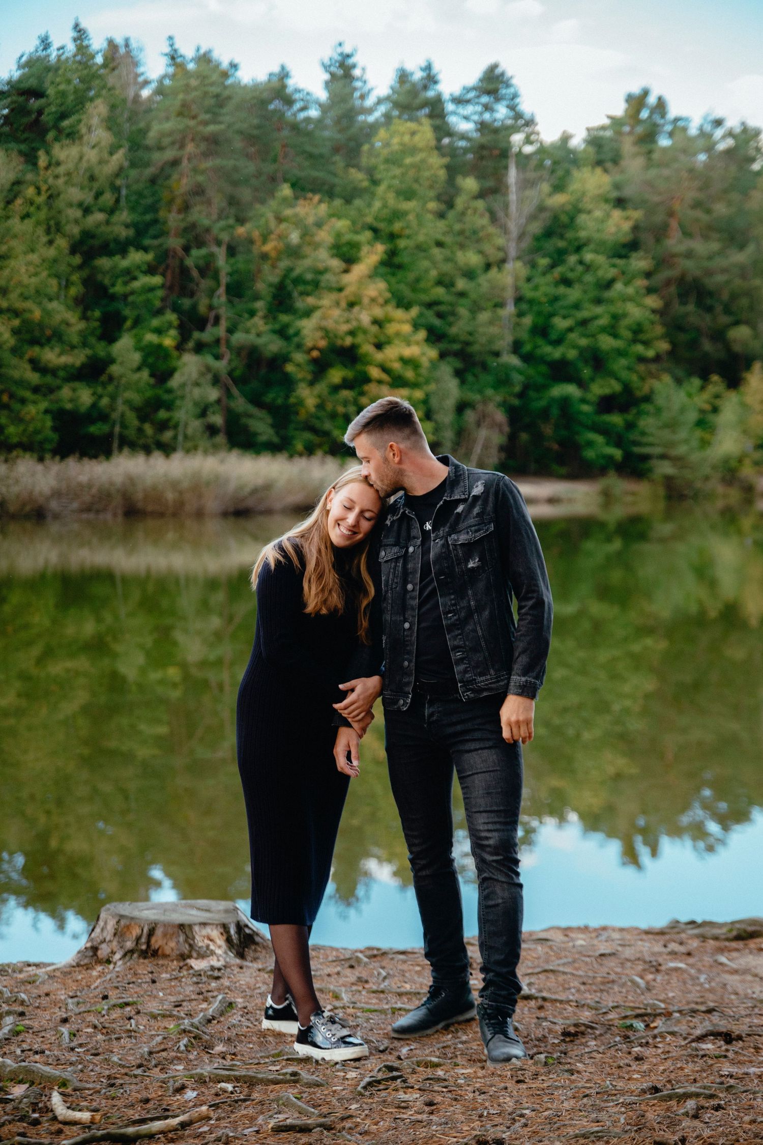 Couple in casual attire poses playfully by a lakeside forest setting.