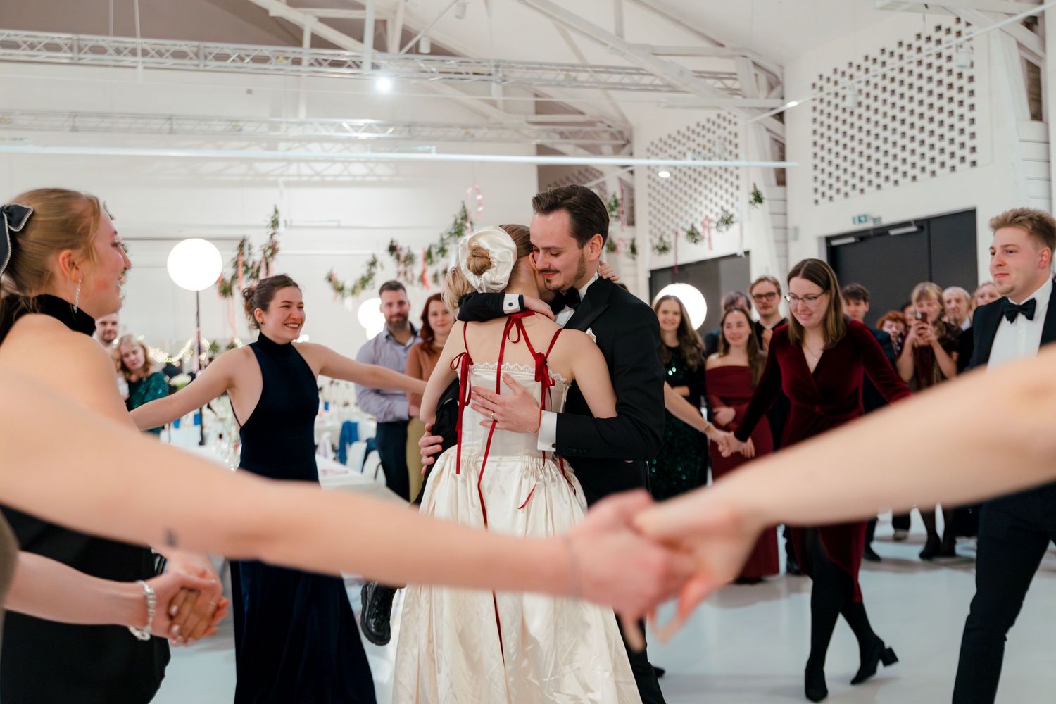Wedding guests dance in pairs across a bright indoor venue space.