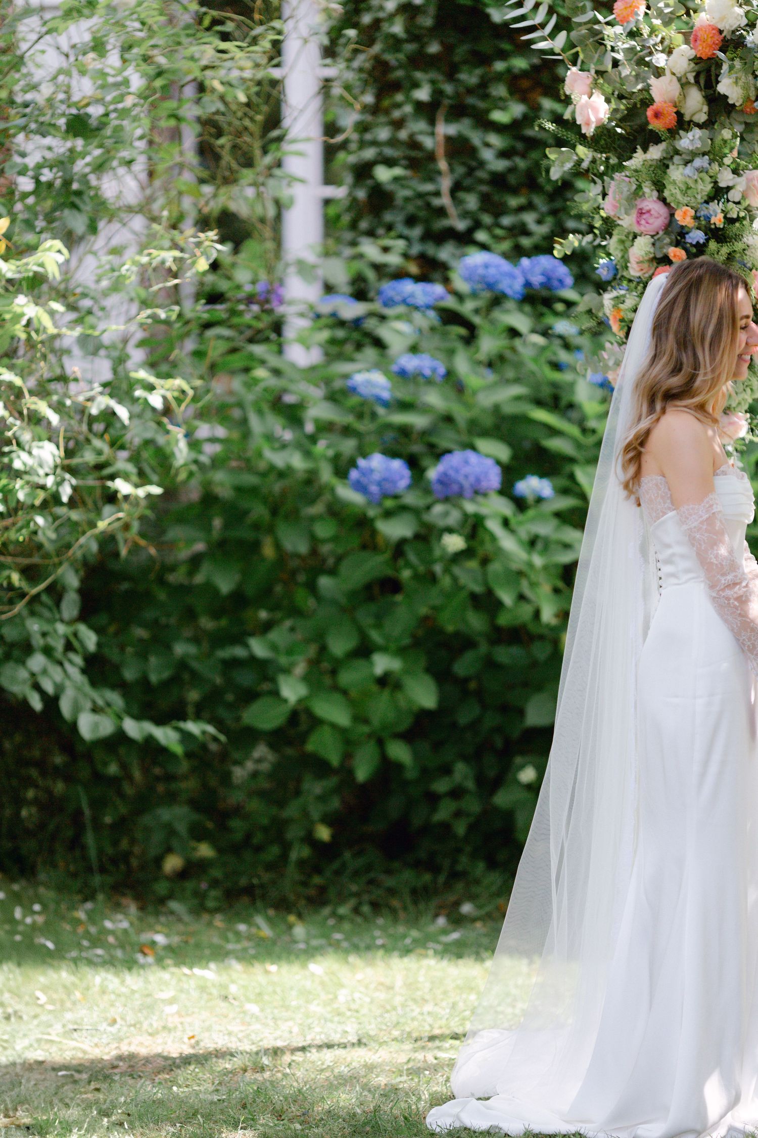 Une mariée élégante en robe en dentelle blanche avec voile se tient sous une arche florale colorée entourée d'hortensias.