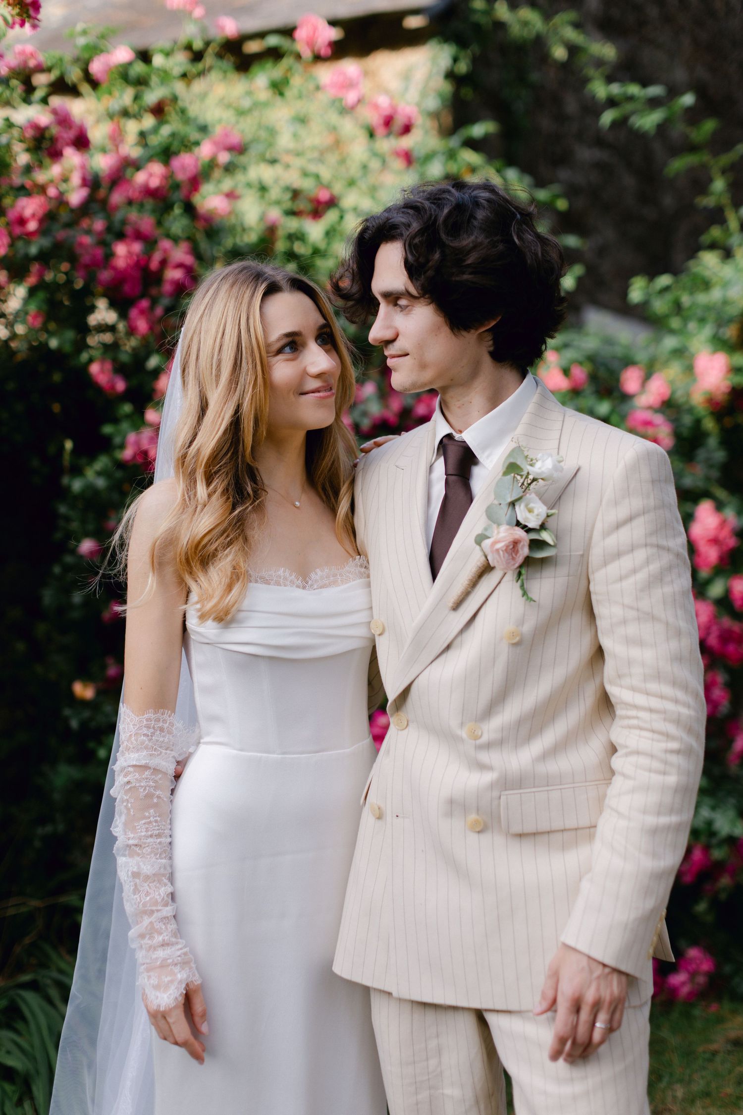 Mariée en robe blanche avec gants en dentelle et marié en costume à rayures beiges avec boutonnière florale pose devant des roses.