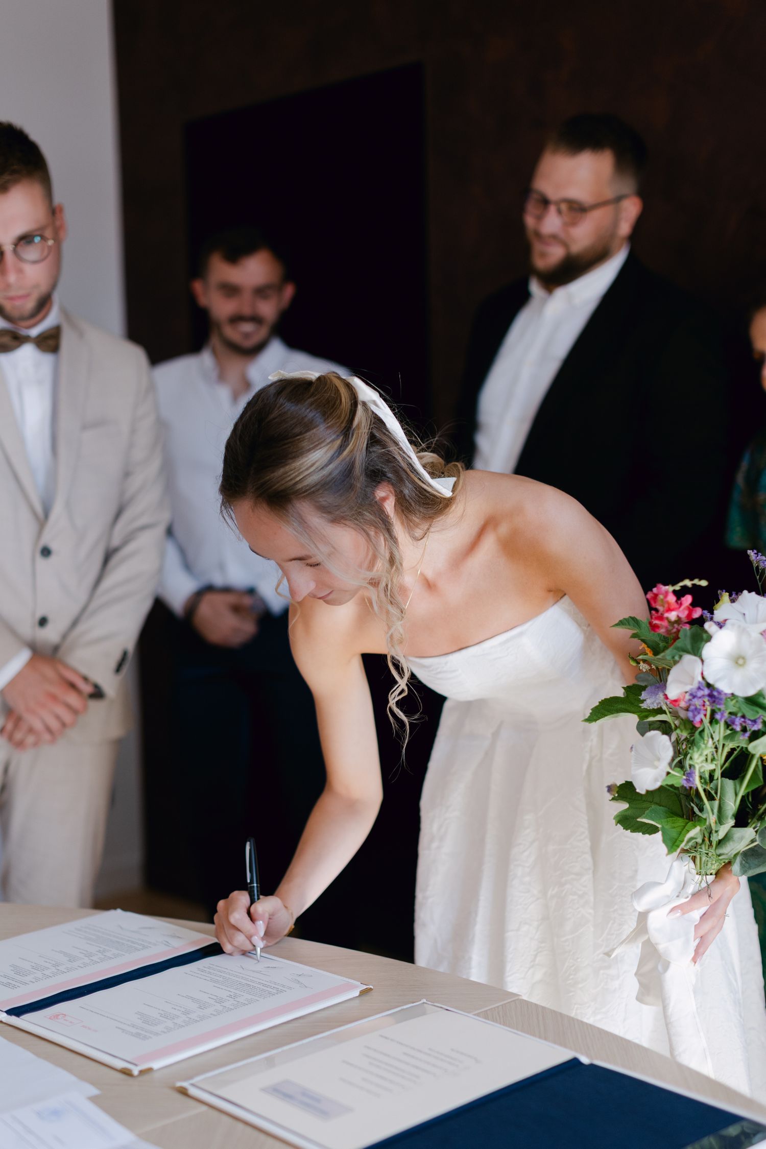 Mariée en robe blanche sans bretelles signant des documents de mariage lors d'une cérémonie civile avec des invités qui regardent.