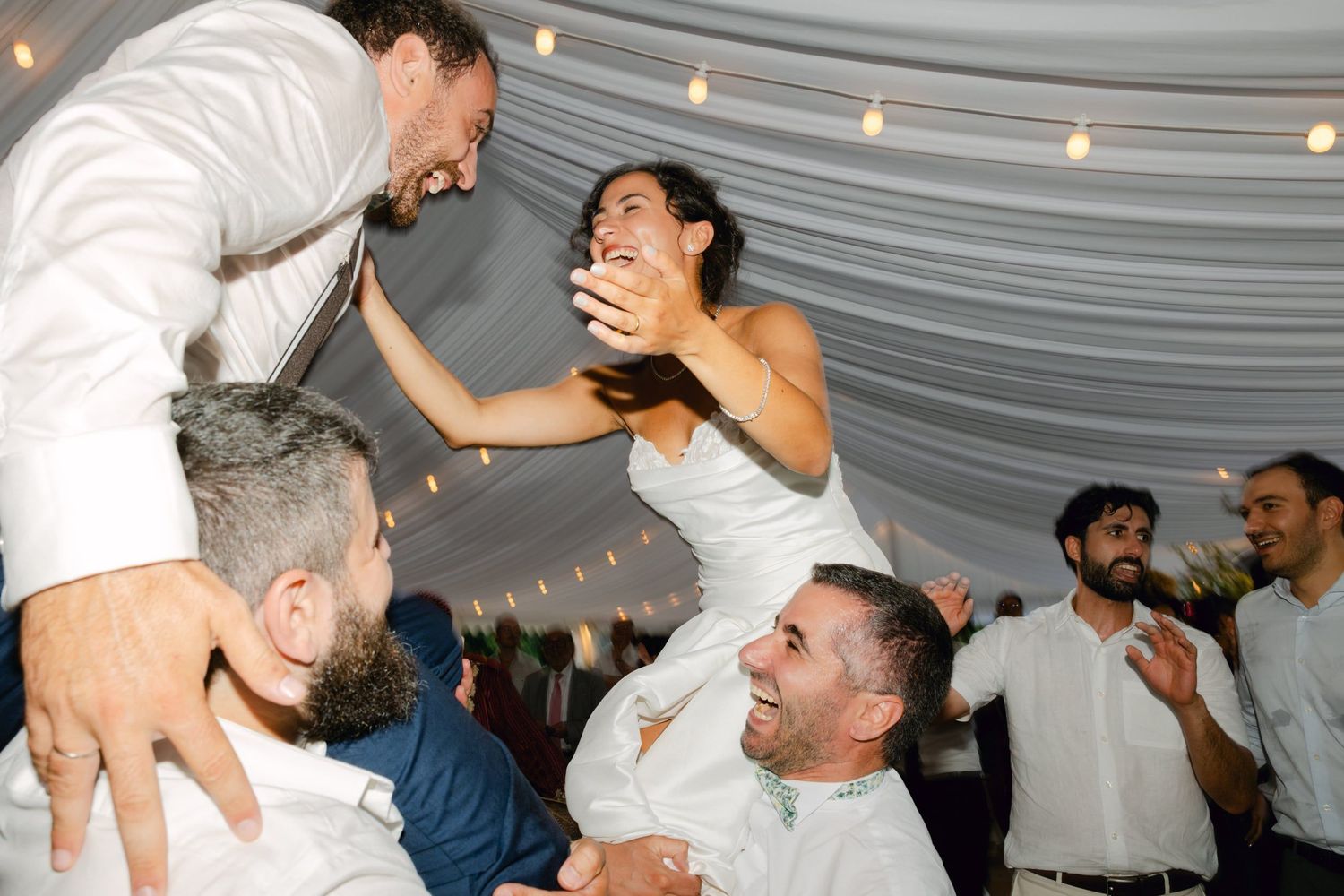 Bride lifted on shoulders by guests during joyful wedding reception celebration under a tent with string lights.