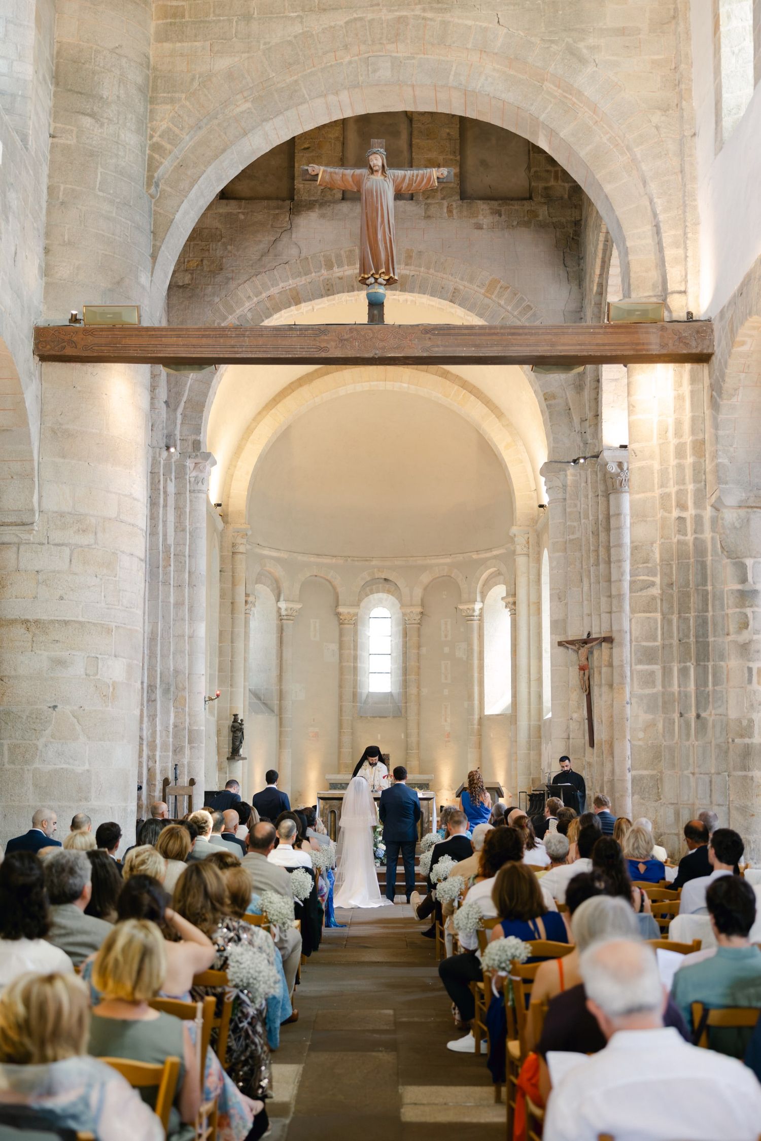 Wedding ceremony in a Romanesque stone church with guests seated in pews and a crucifix above the altar.