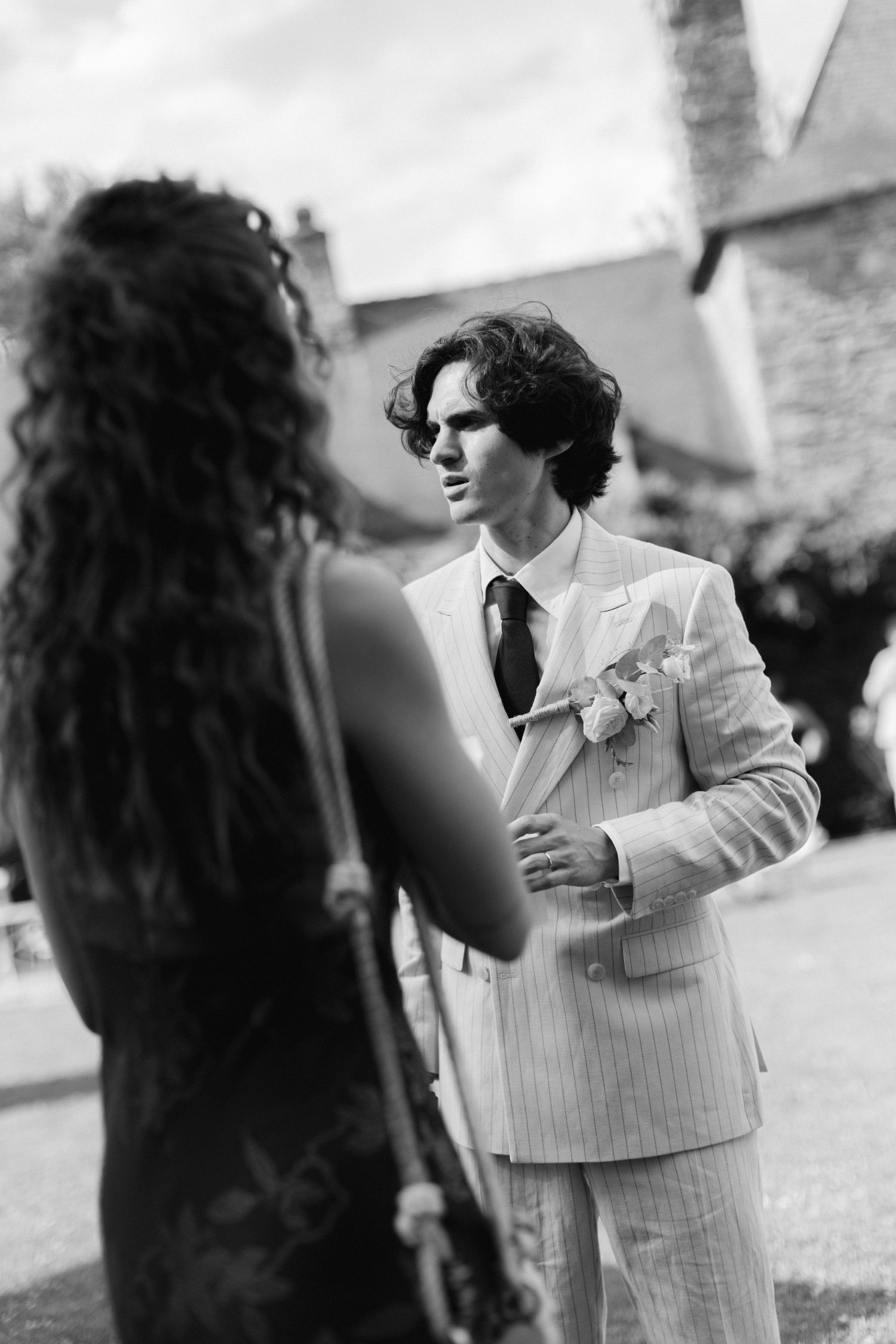 Photo en noir et blanc d'un marié en costume à rayures fines parlant à un invité aux cheveux bouclés à l'extérieur.