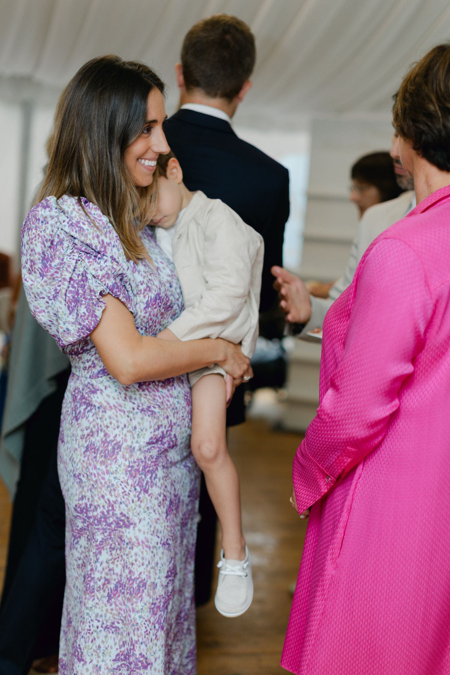 Invité de mariage en robe à fleurs violettes tenant un jeune enfant tout en socialisant à la réception.