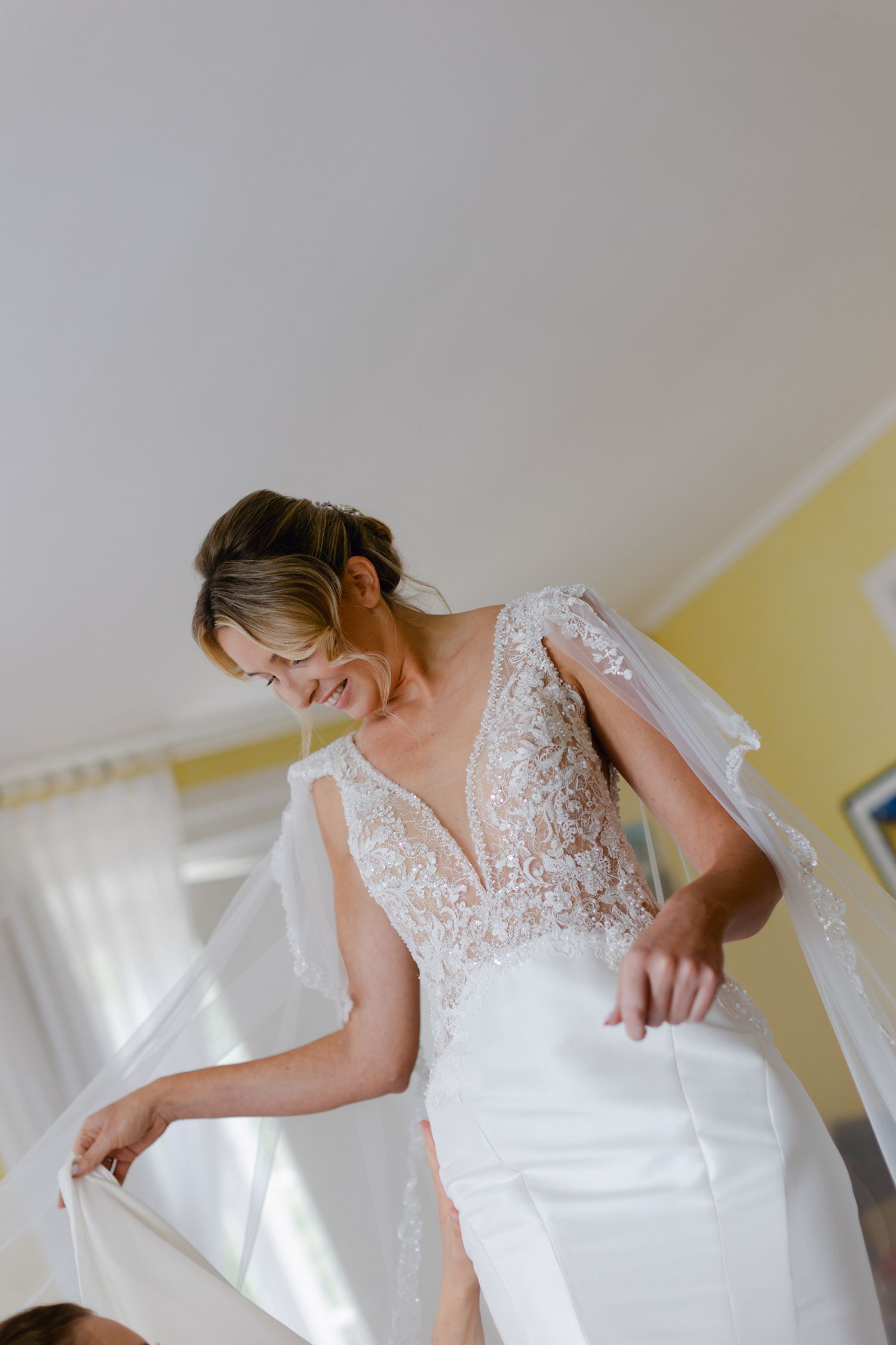 Mariée souriante en robe de mariée en dentelle à col en V profond tenant son voile pendant les préparatifs de la mariée.