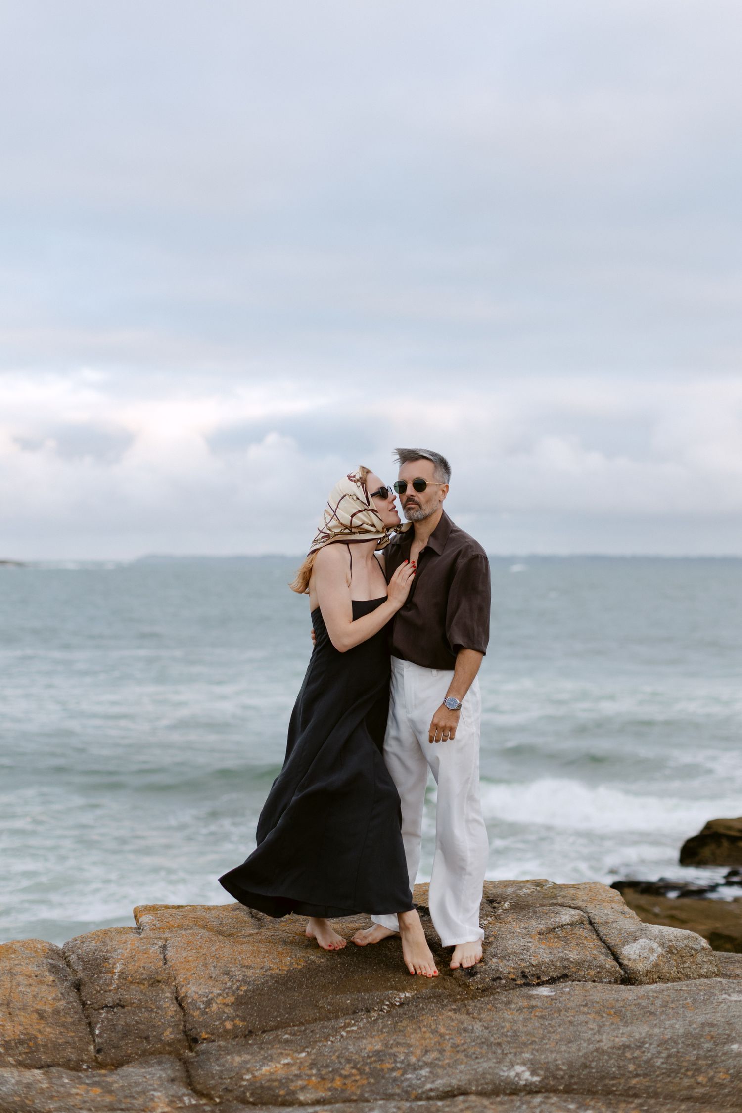 Couple debout sur les rochers côtiers au bord de l'océan, femme en robe noire et homme en pantalon blanc et chemise marron.