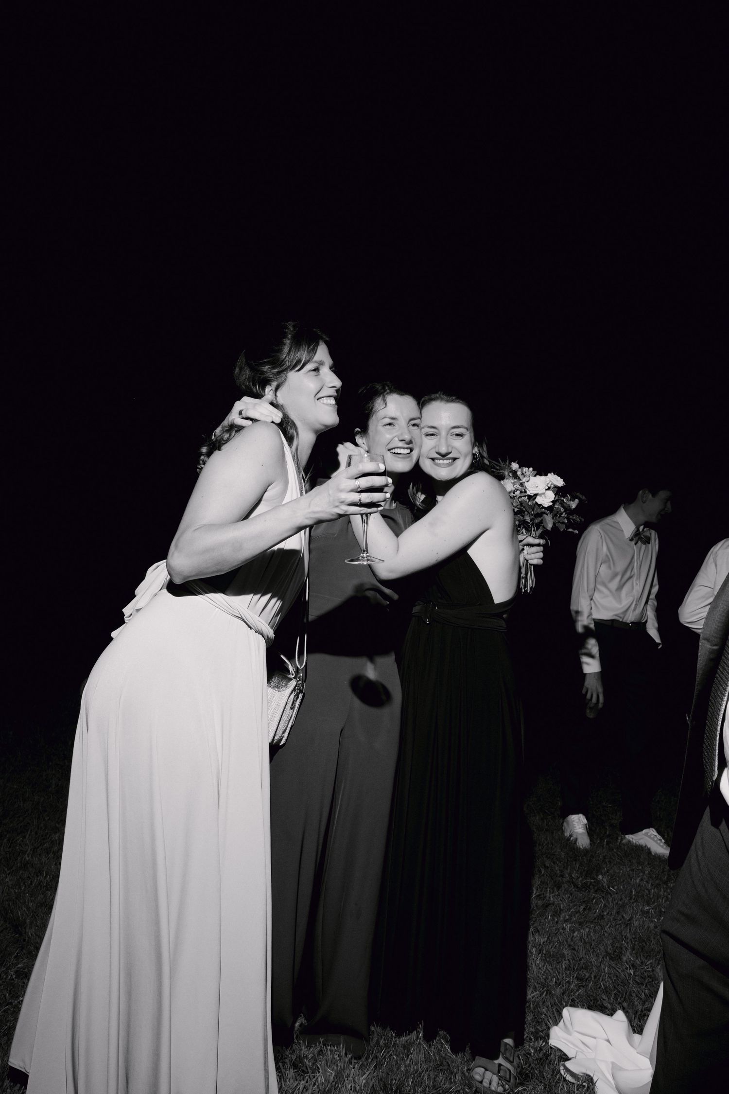 Photo en noir et blanc de trois femmes en robes formelles souriantes et s'embrassant lors d'une réception de mariage en soirée en plein air.
