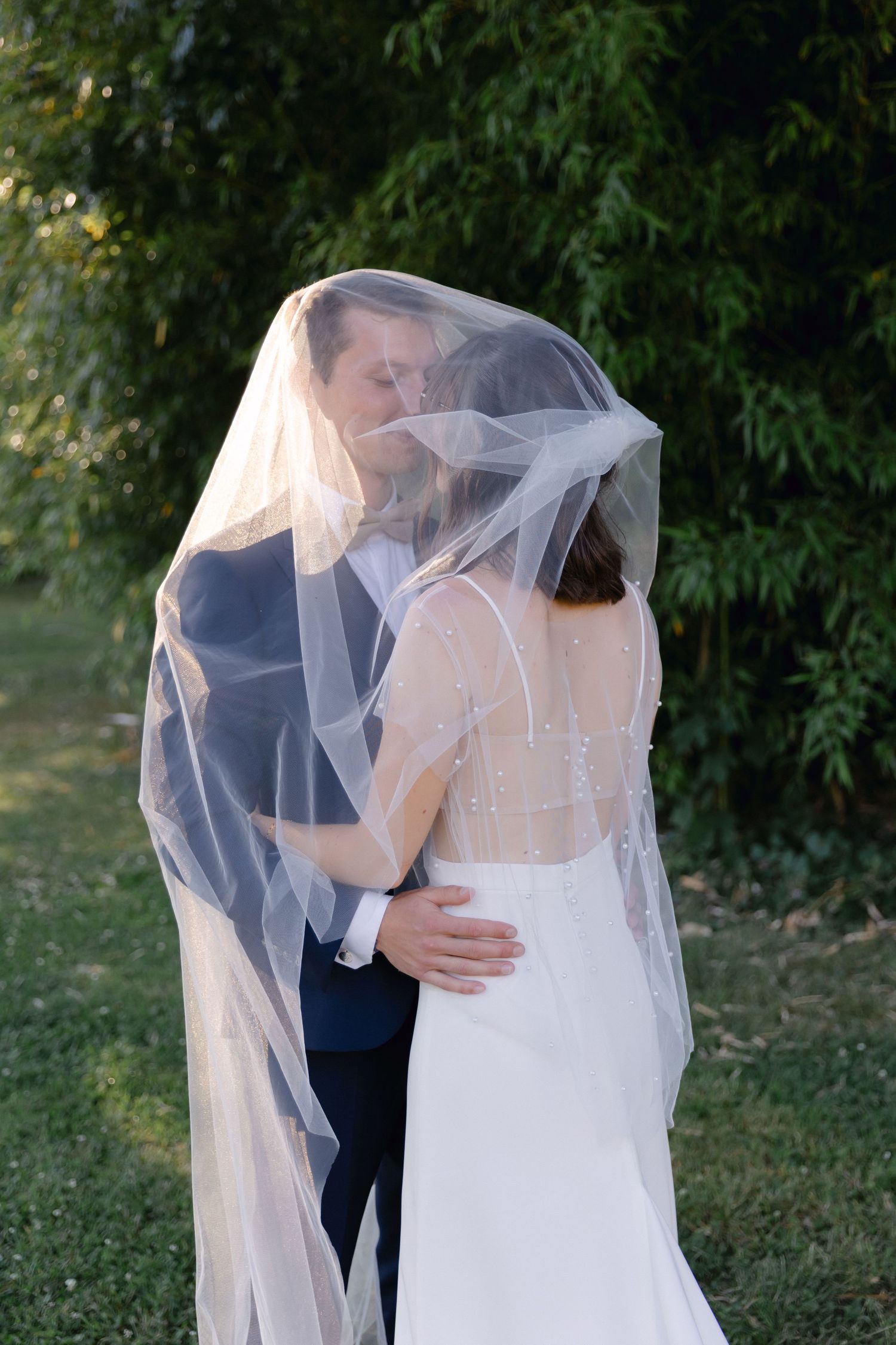 La mariée et le marié partagent un baiser romantique sous un voile blanc fluide à l'extérieur entouré d'arbres verts luxuriants.