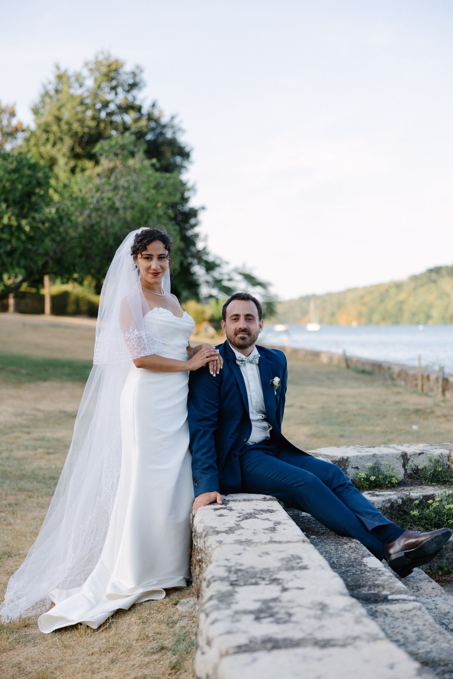 Bride in white gown and veil poses with groom in navy suit on stone wall by waterfront.