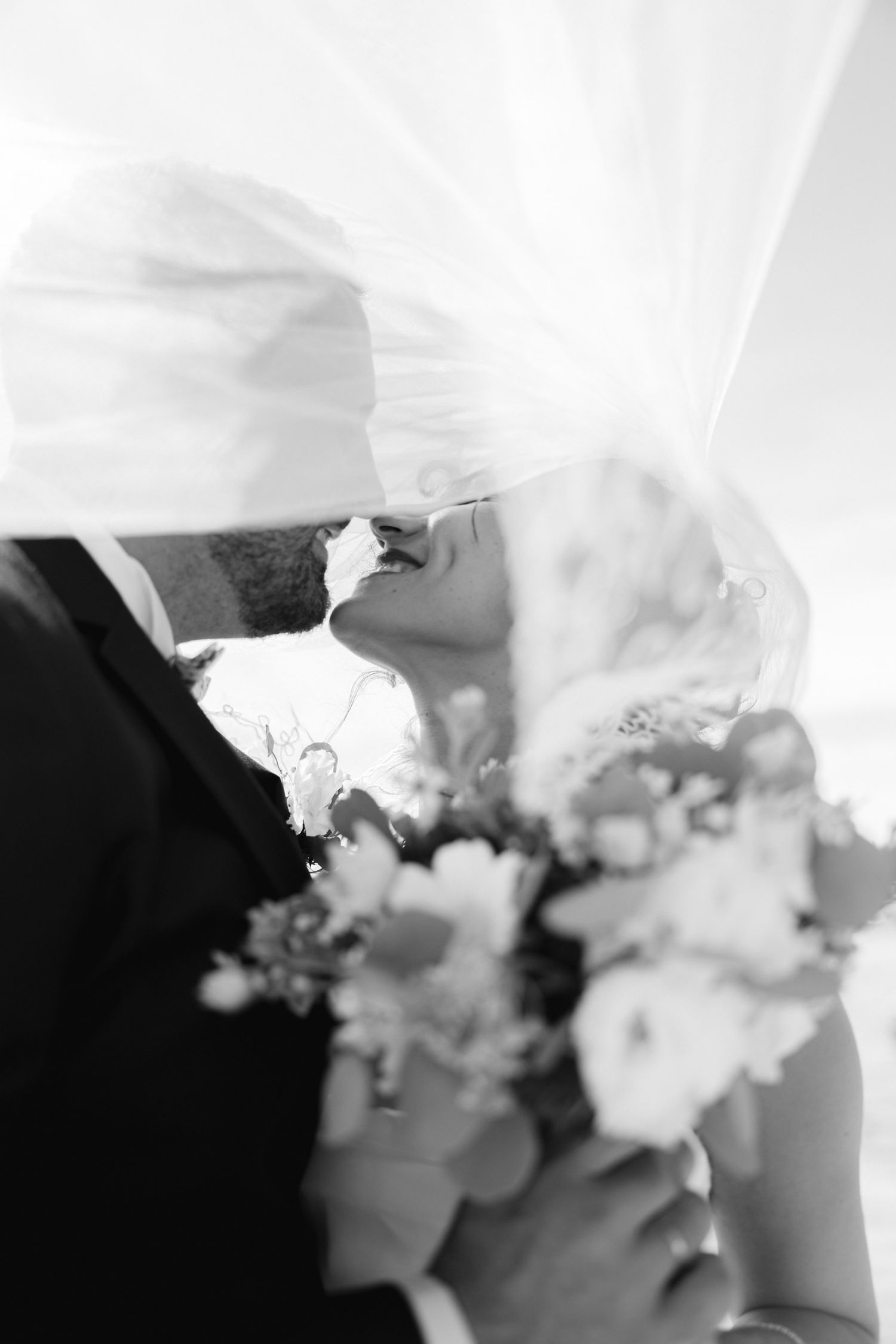Black and white photo of bride and groom kissing under veil, bride holding floral bouquet.