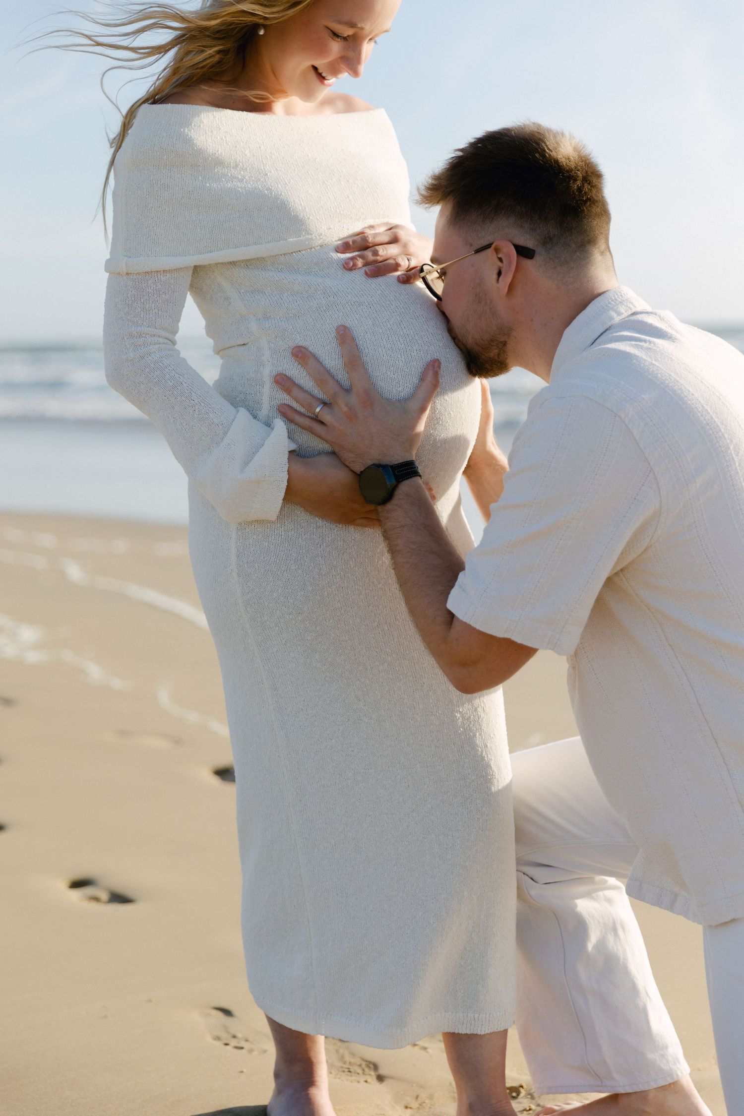 Un homme s'agenouille pour embrasser le ventre d'une femme enceinte sur une plage ensoleillée, tous deux vêtus de tenues blanches.