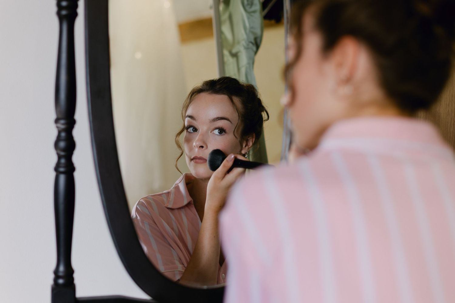 Demoiselle d'honneur en chemise rayée rose se maquillant avec un pinceau tout en regardant dans un miroir orné.