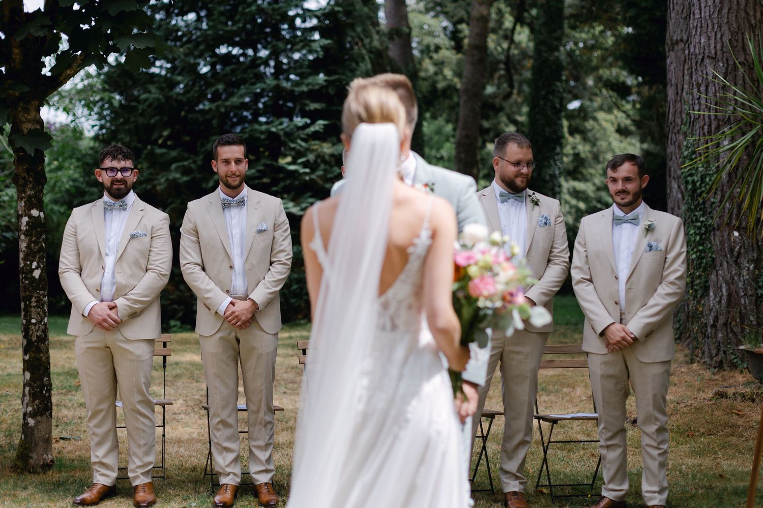 La mariée en robe blanche se dirige vers les garçons d'honneur en costume beige lors d'une cérémonie de mariage en plein air.