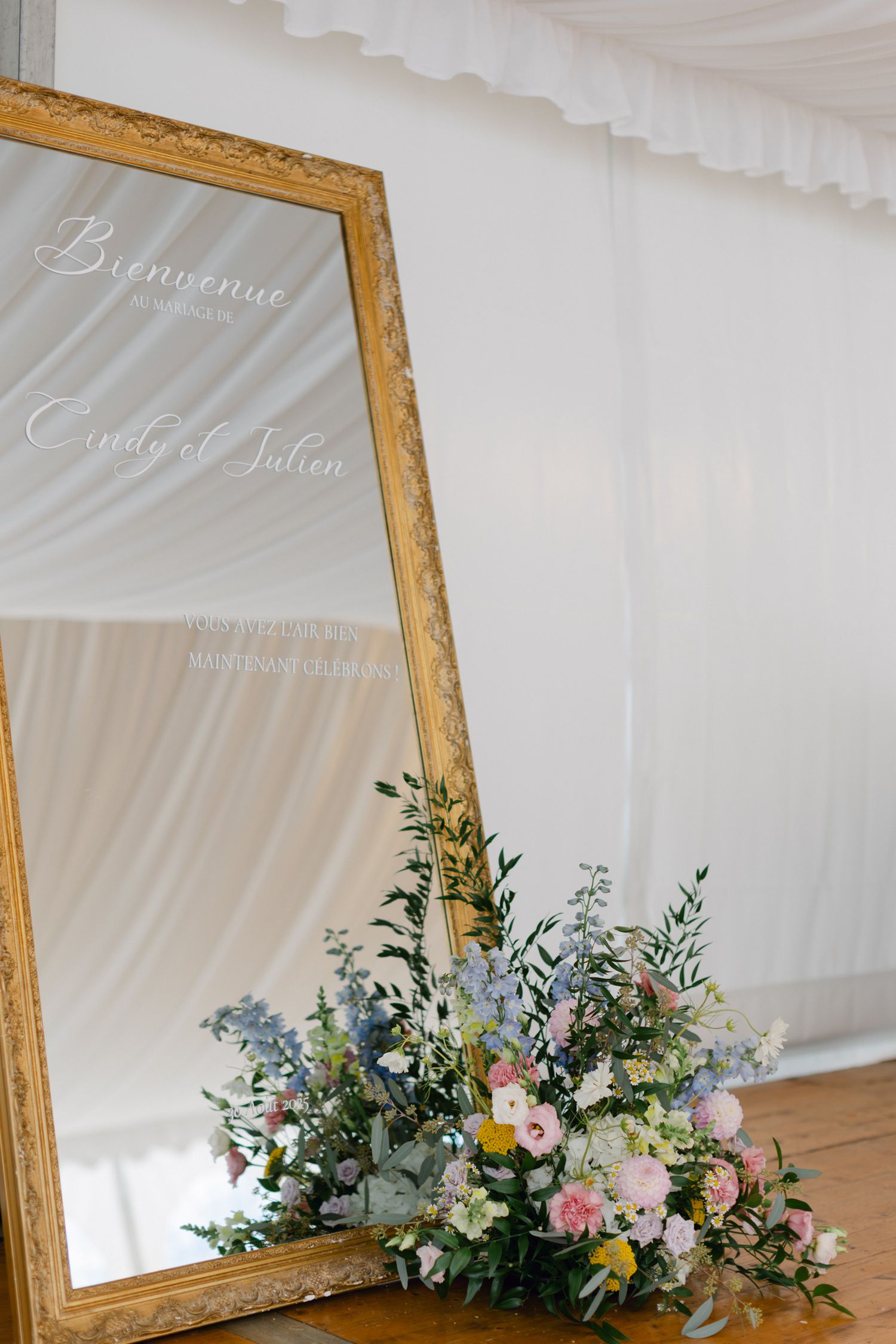 Élégant miroir encadré d'or avec message de bienvenue de mariage et arrangement floral pastel à la base.