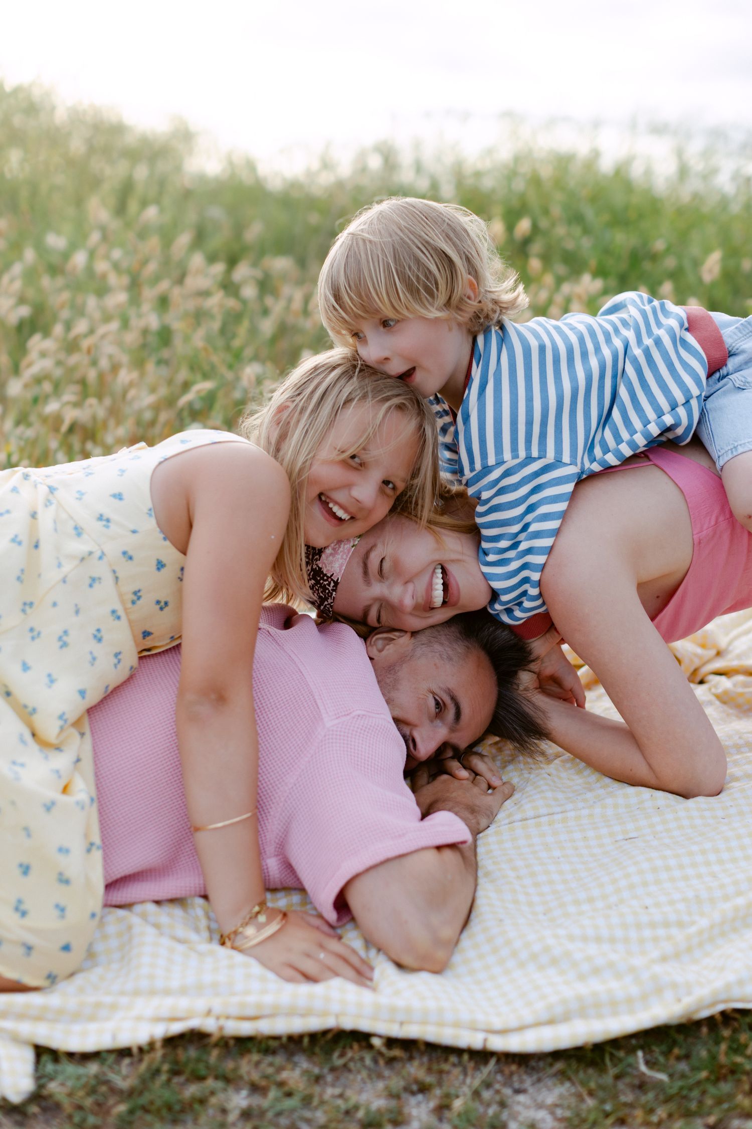 Famille heureuse riant ensemble sur une couverture à l'extérieur, mère et enfant empilant sur papa dans un champ herbeux.