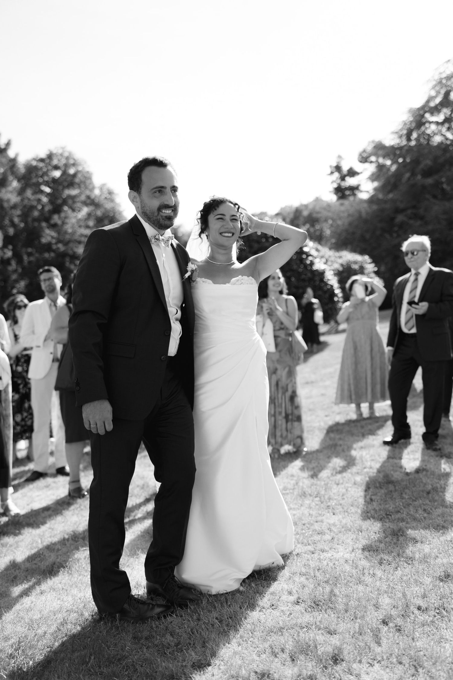 Black and white photo of smiling bride and groom walking outdoors at wedding reception with guests in background.