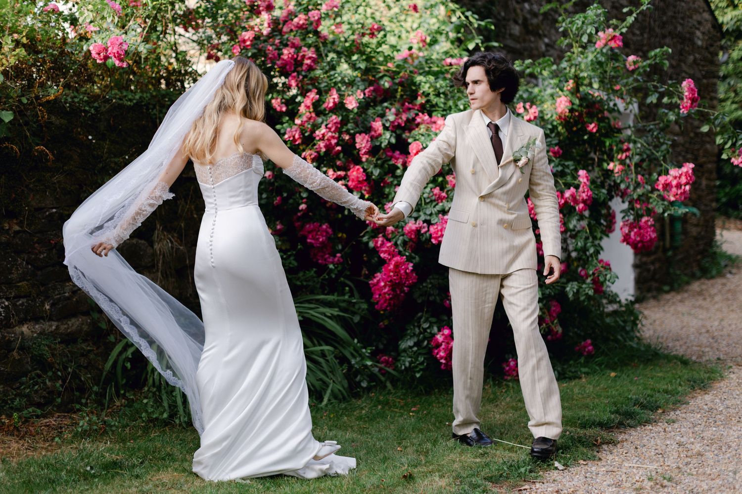 La mariée en robe blanche fluide et voile tient la main du marié près de roses roses vibrantes dans un cadre de mariage dans un jardin.
