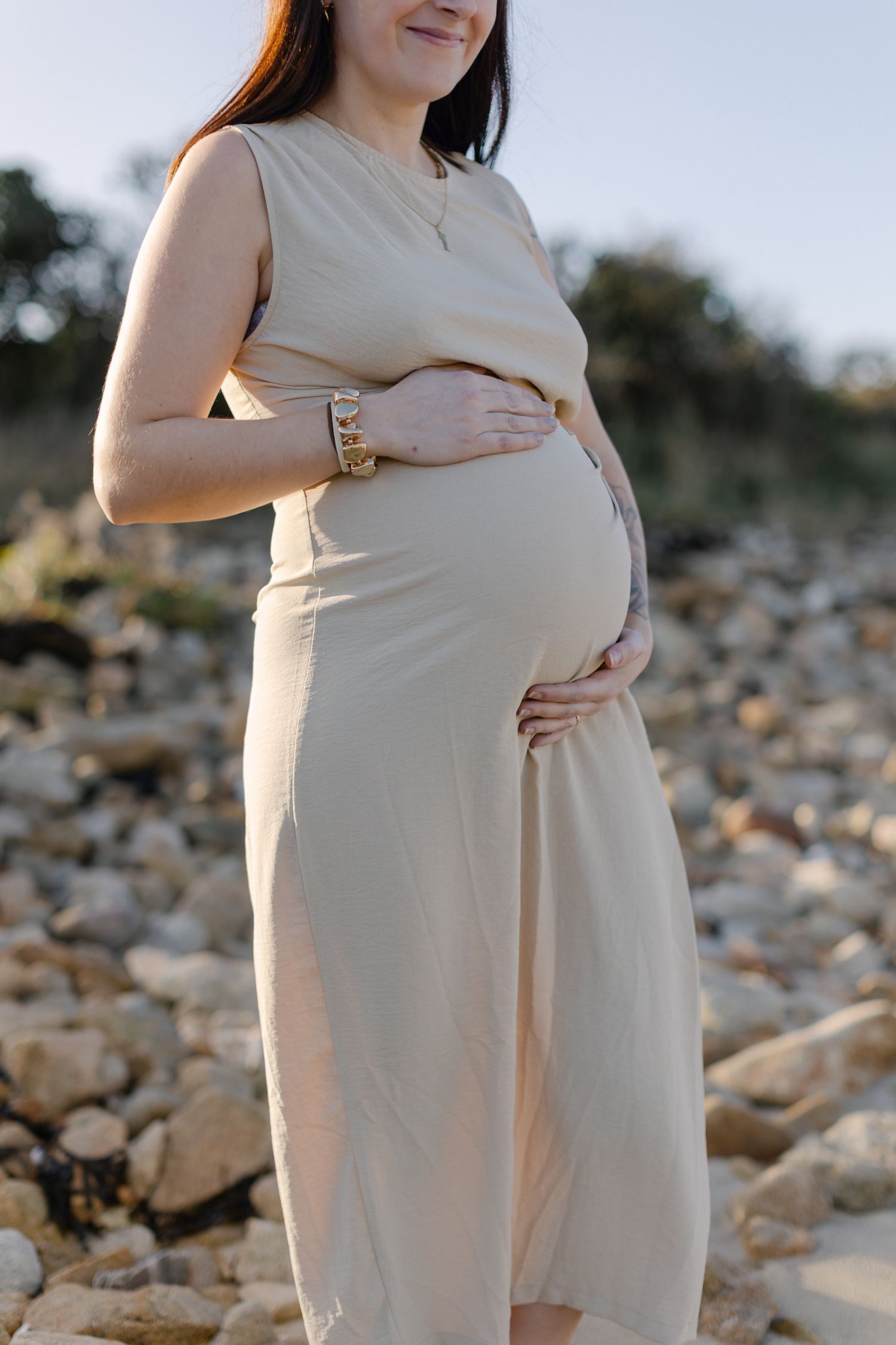 Gros plan d'une femme enceinte en robe beige berçant le ventre de bébé avec les deux mains sur une plage rocheuse.