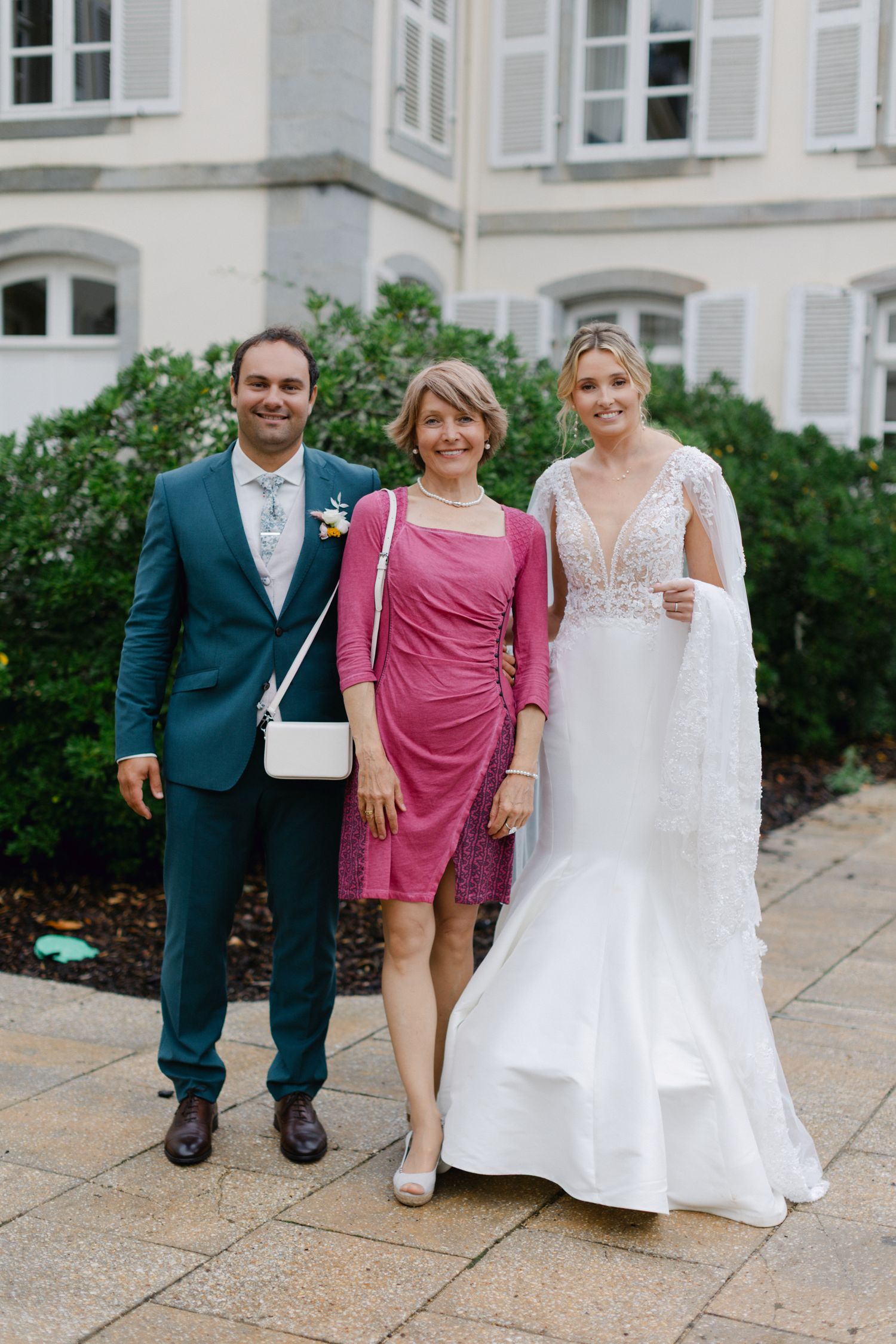 La mariée en robe de dentelle et le marié en costume sarcelle posant avec une femme en robe rose à l'extérieur du lieu du mariage.