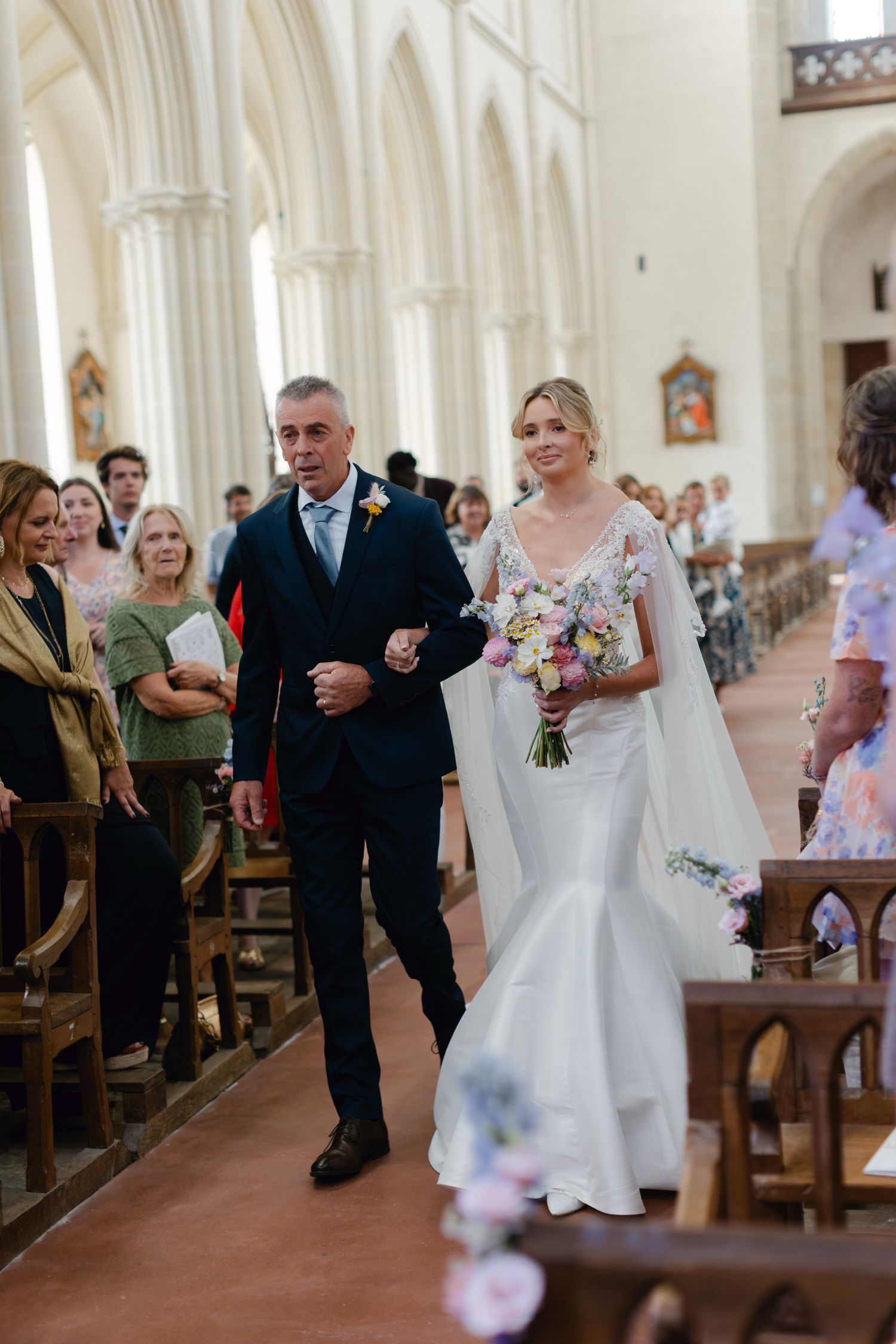 La mariée en robe de sirène blanche avec bouquet marche dans l'allée de l'église escortée par son père pendant le mariage.