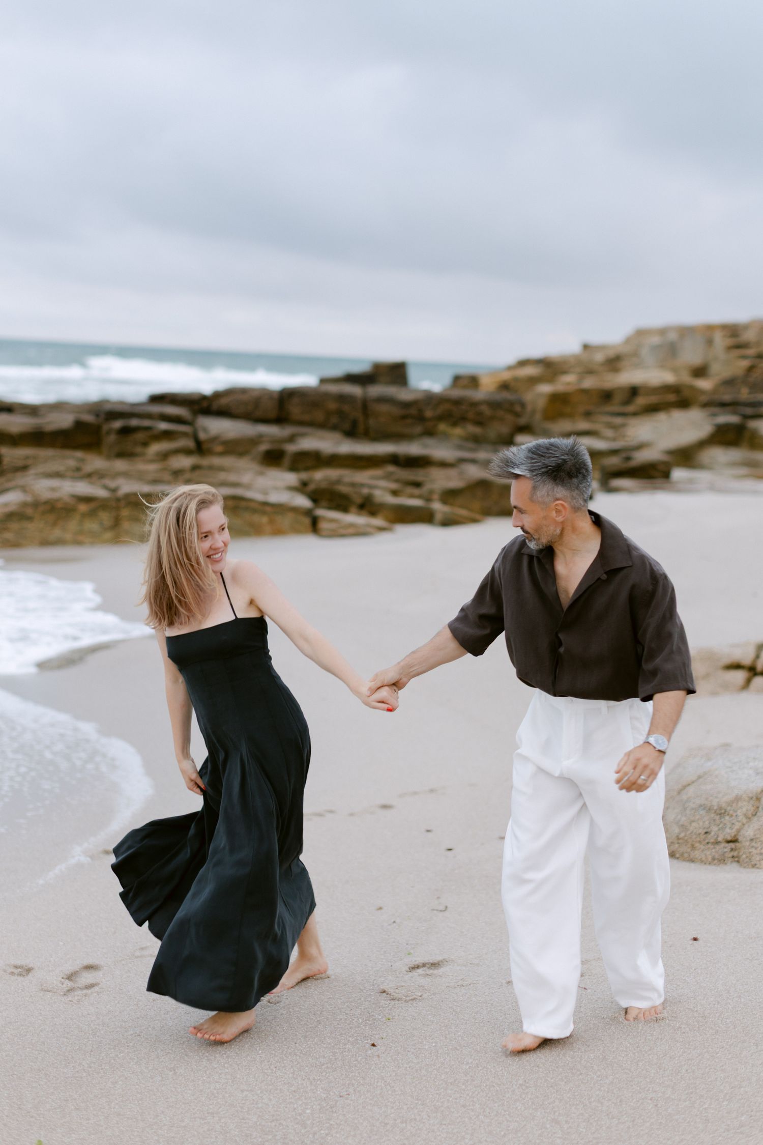 Couple dansant pieds nus sur une plage de sable, femme en robe noire et homme en pantalon blanc se tenant la main près du rivage rocheux.