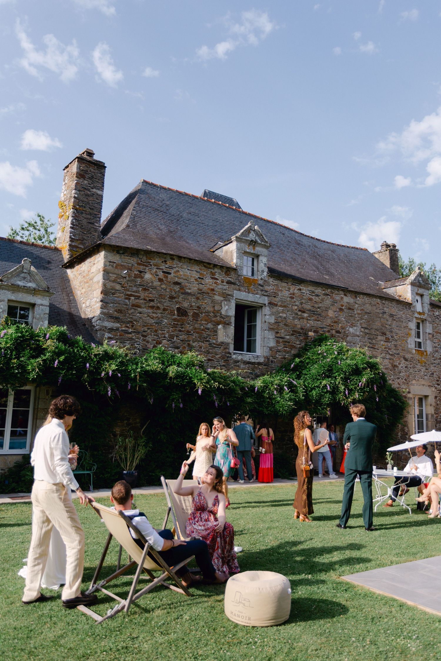 Les invités au mariage se détendent sur des chaises de jardin à l'extérieur d'une maison de campagne rustique en pierre française.