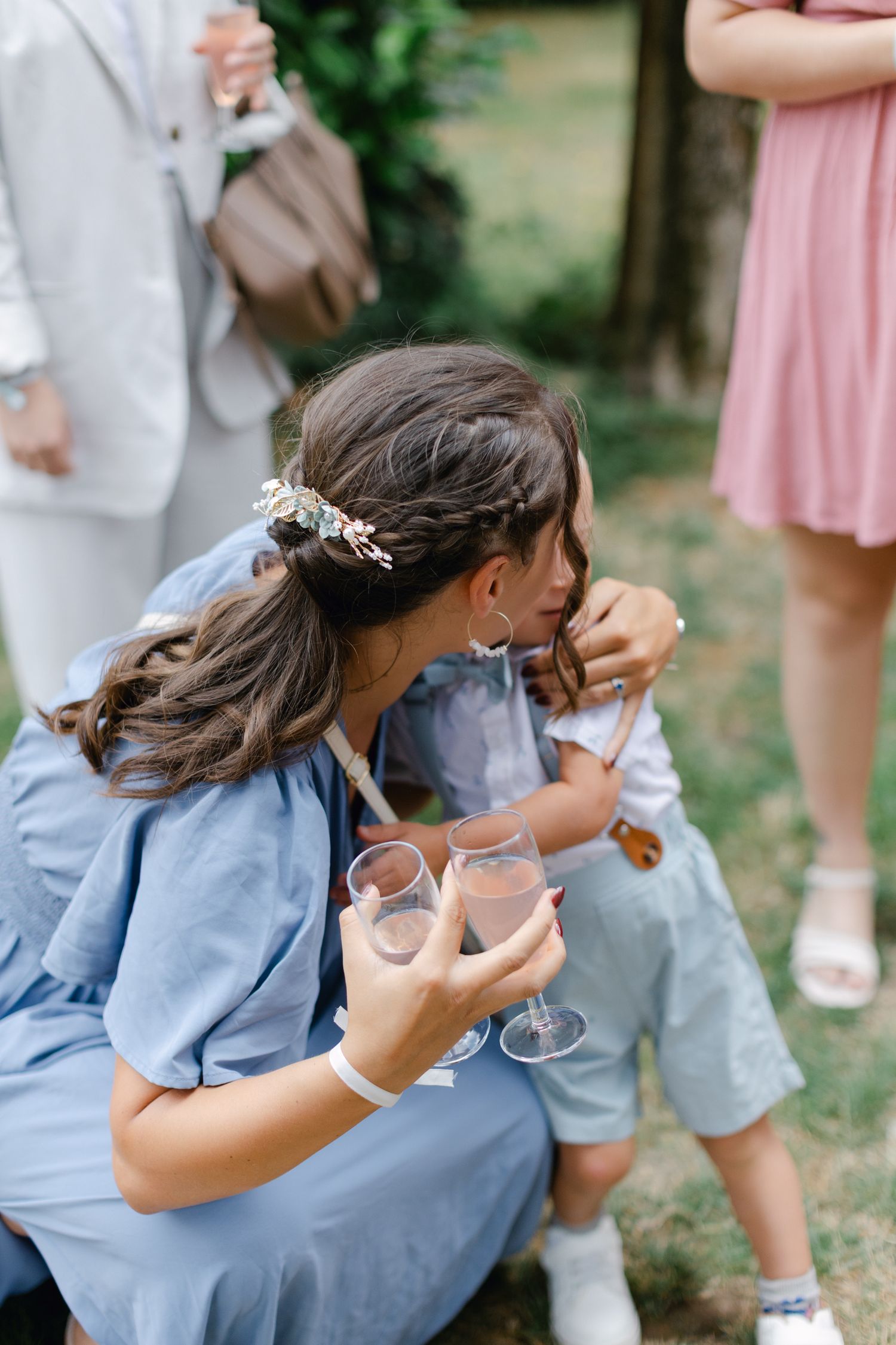 Femme en robe bleue accroupie pour interagir avec un jeune enfant lors d'une réception de mariage en plein air tenant du champagne.