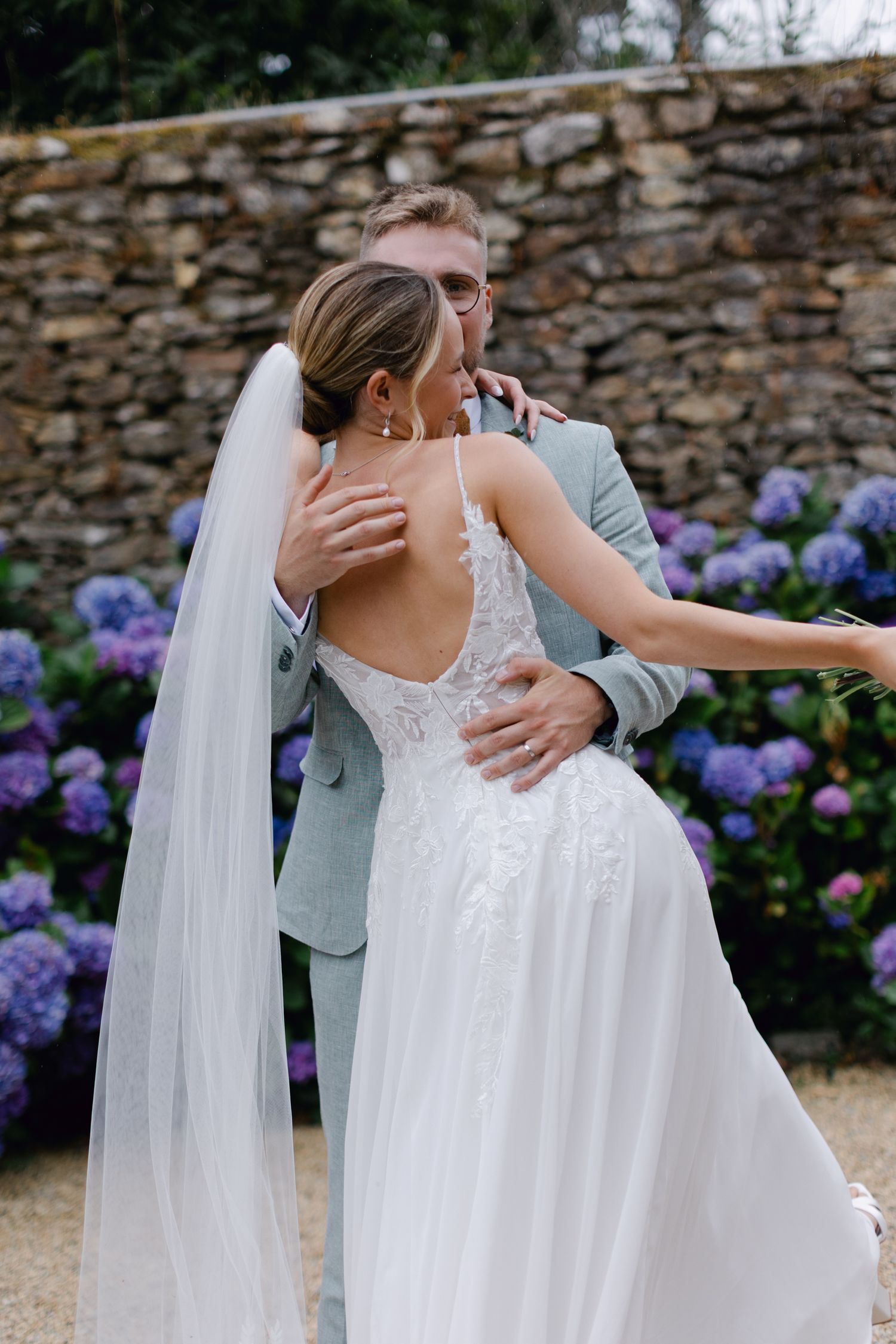 Le marié embrasse la mariée par derrière montrant une robe de mariée en dentelle à dos ouvert avec un voile près des hortensias.