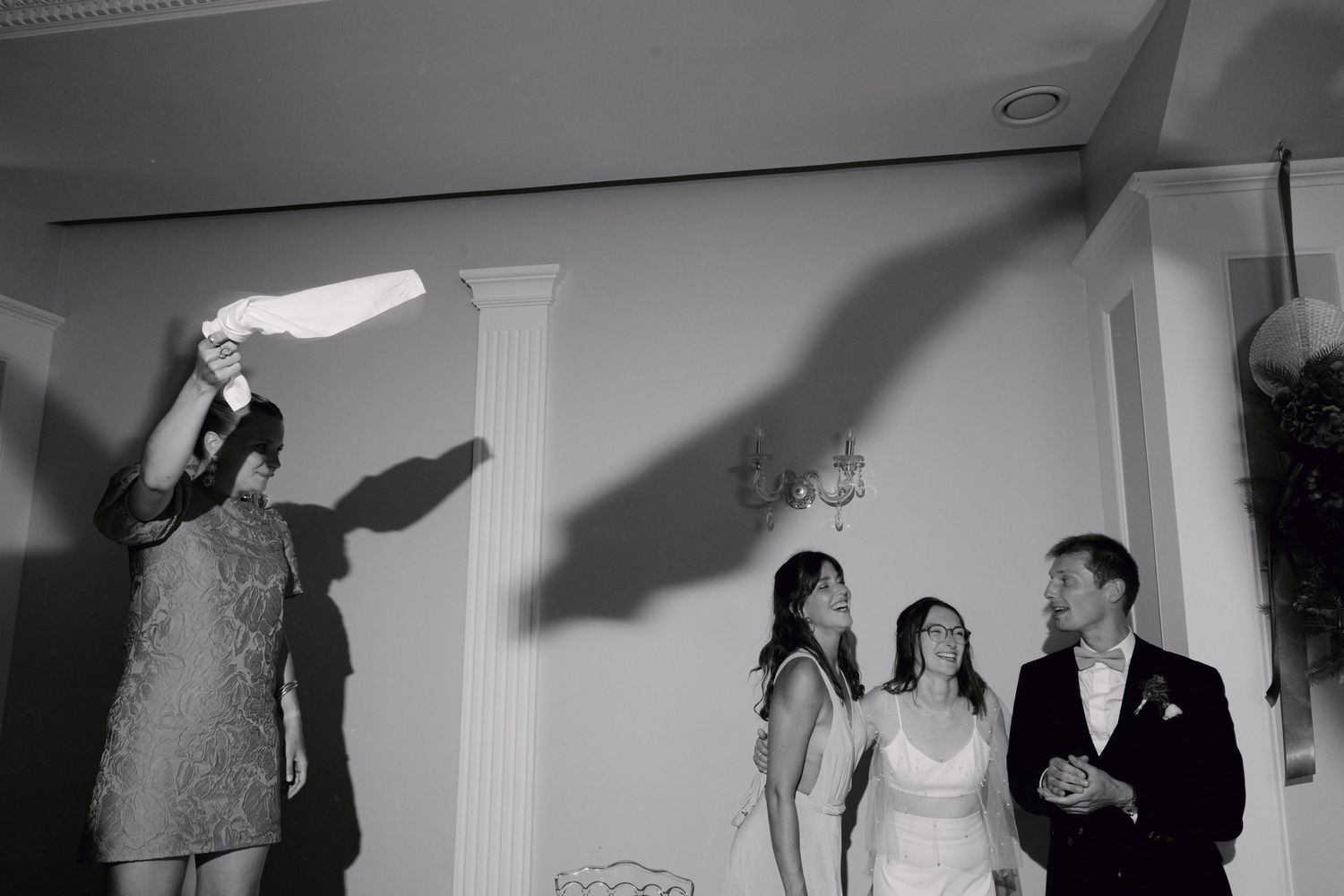 Photo de mariage en noir et blanc de la mariée et du marié avec des invités célébrant à l'intérieur près des colonnes blanches.