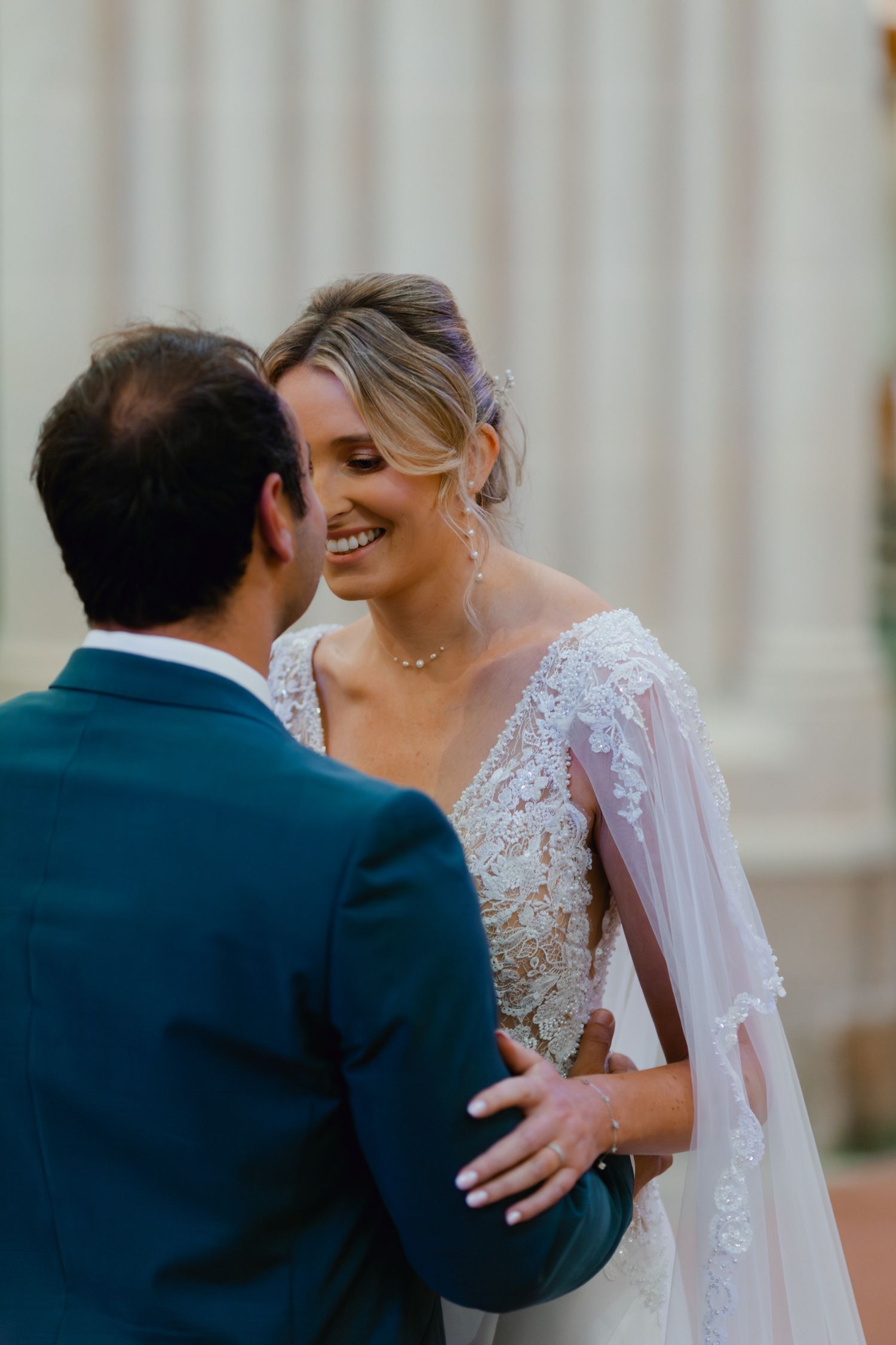 La mariée souriante en robe de mariée en dentelle avec voile de cape partage un moment intime avec le marié en costume sarcelle.