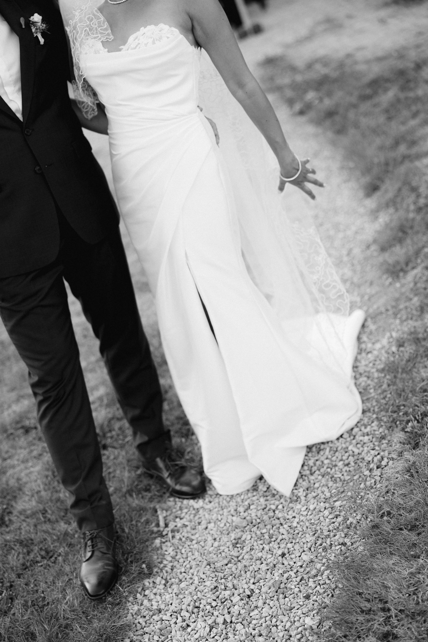 Black and white photo of bride in white gown and groom in dark suit walking on gravel path at wedding.