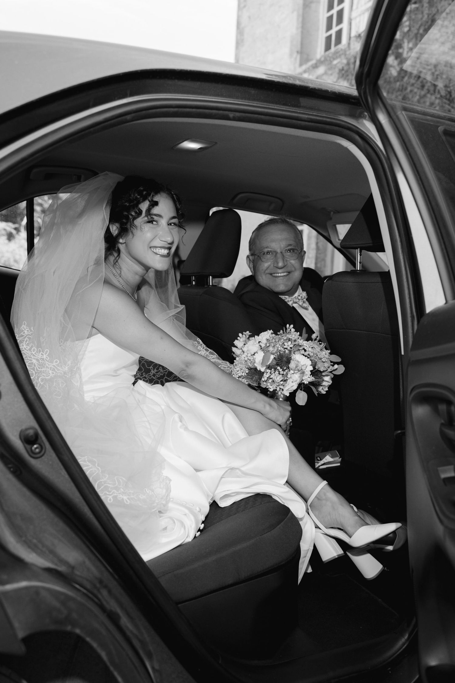 Black and white photo of a smiling bride in white dress holding bouquet sitting in car with older man.