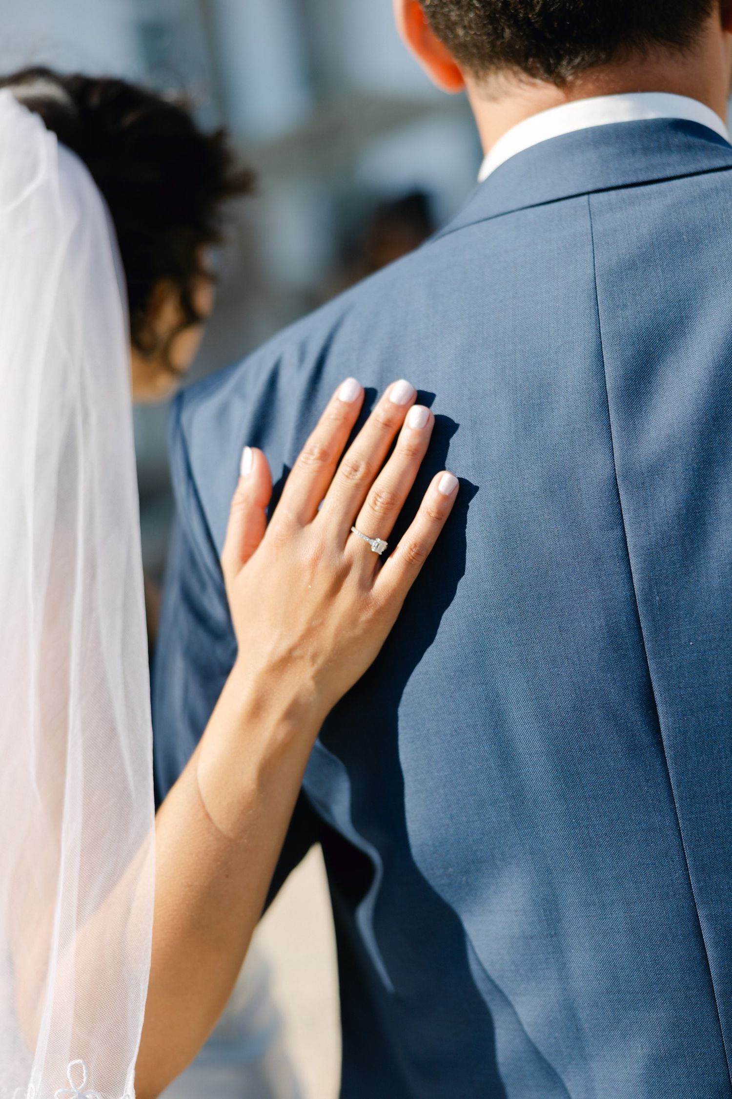 Bride placing her hand on groom's blue suit jacket, showing off her engagement ring and wedding veil.