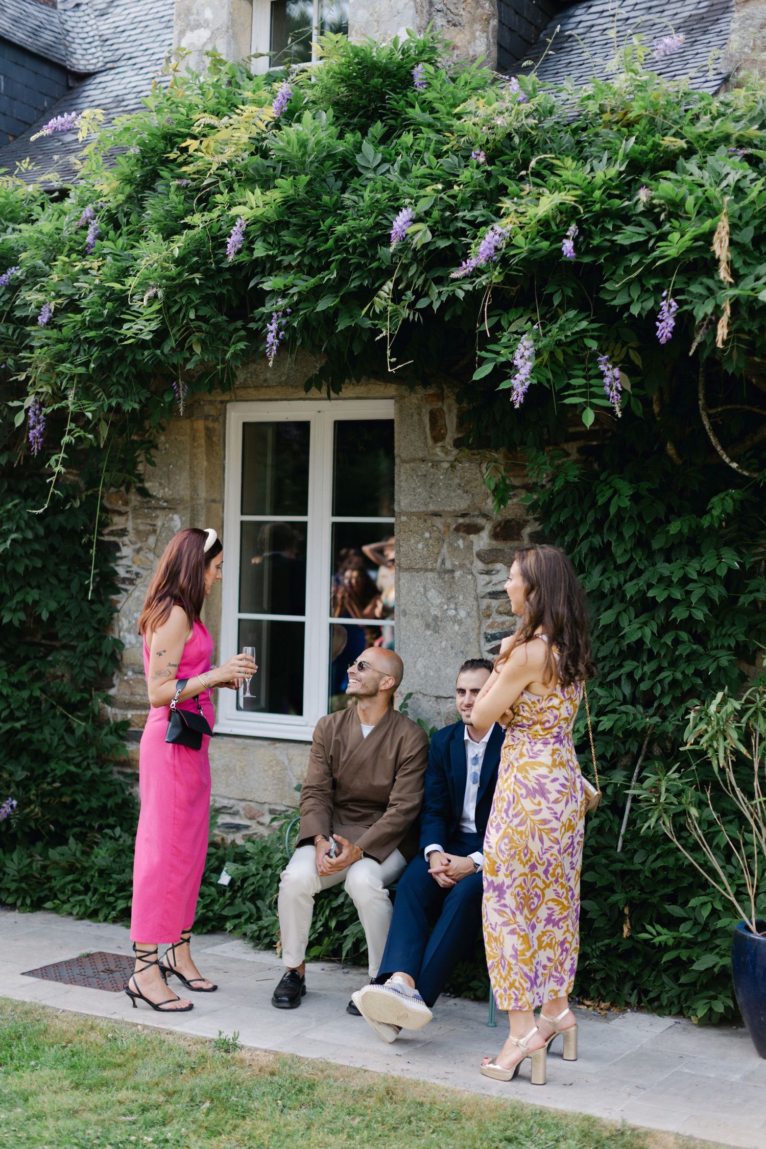 Les invités au mariage en tenue colorée discutent sous une façade de bâtiment en pierre drapée de glycines.