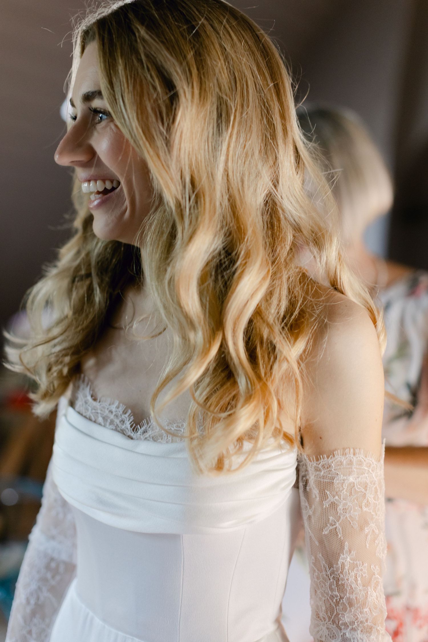 Mariée souriante dans une robe de mariée blanche sans bretelles avec des manches en dentelle et des cheveux ondulés blonds fluides.
