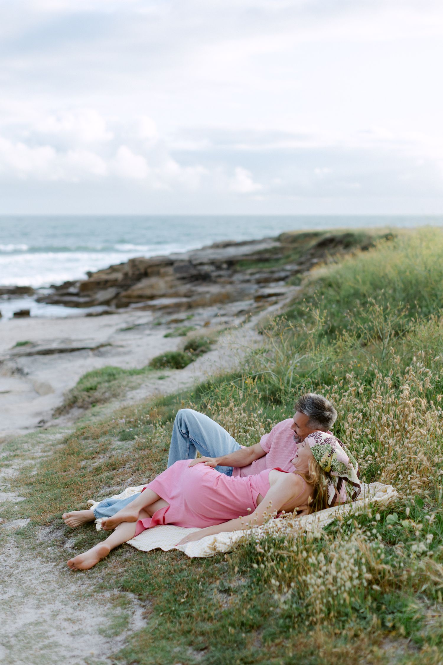 Couple se relaxant sur une couverture près des falaises côtières rocheuses, femme en robe rose, homme en bleu, avec vue sur l'océan.